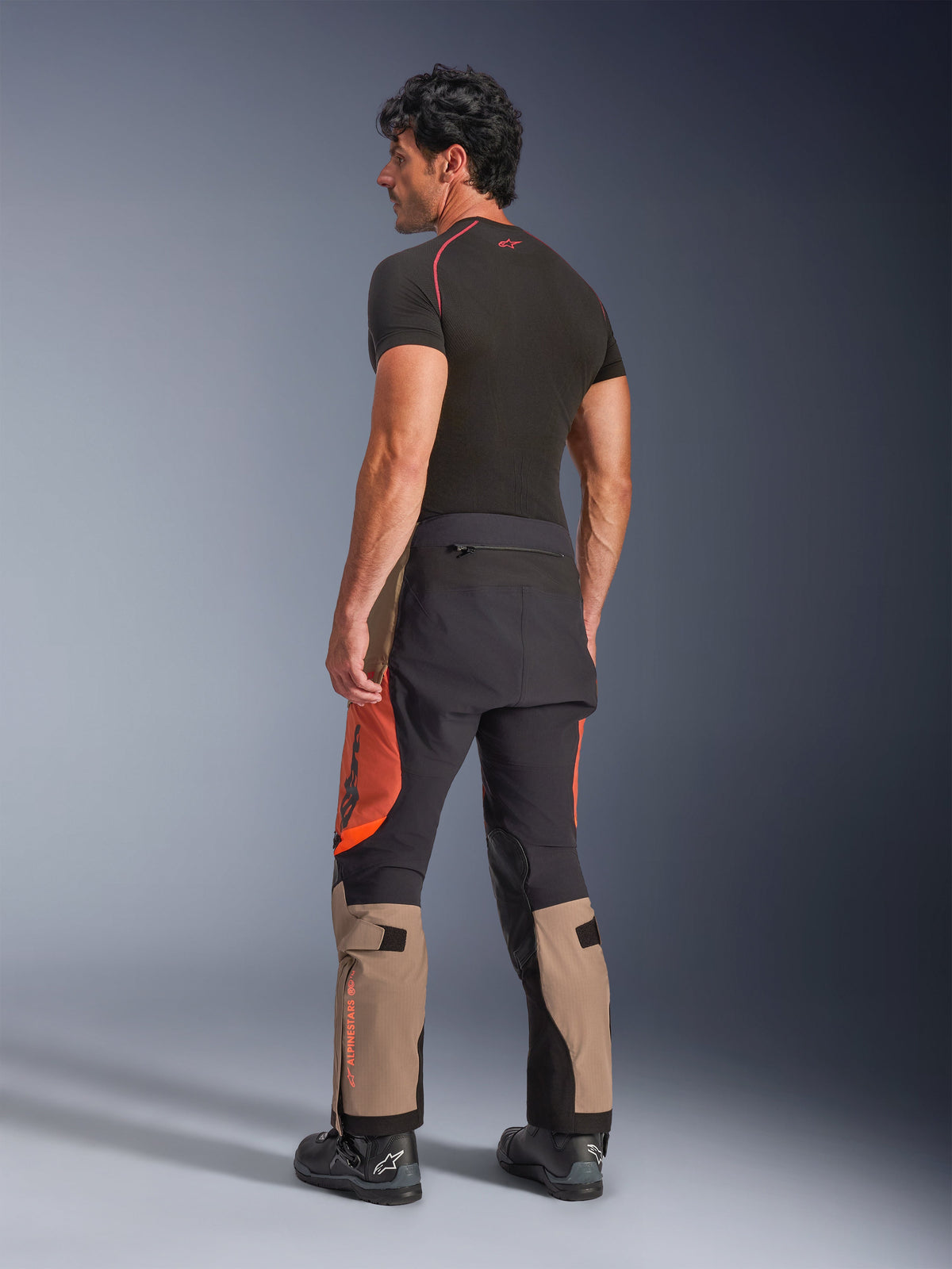 Alpinestars Halo Pro Drystar® XF Laminated Pants in Walnut Rust Black, back view of a male model wearing adventure motorcycle pants with tan, orange, and black panels, styled with a black short-sleeve base layer and black motorcycle boots, highlighting technical outdoor riding gear
