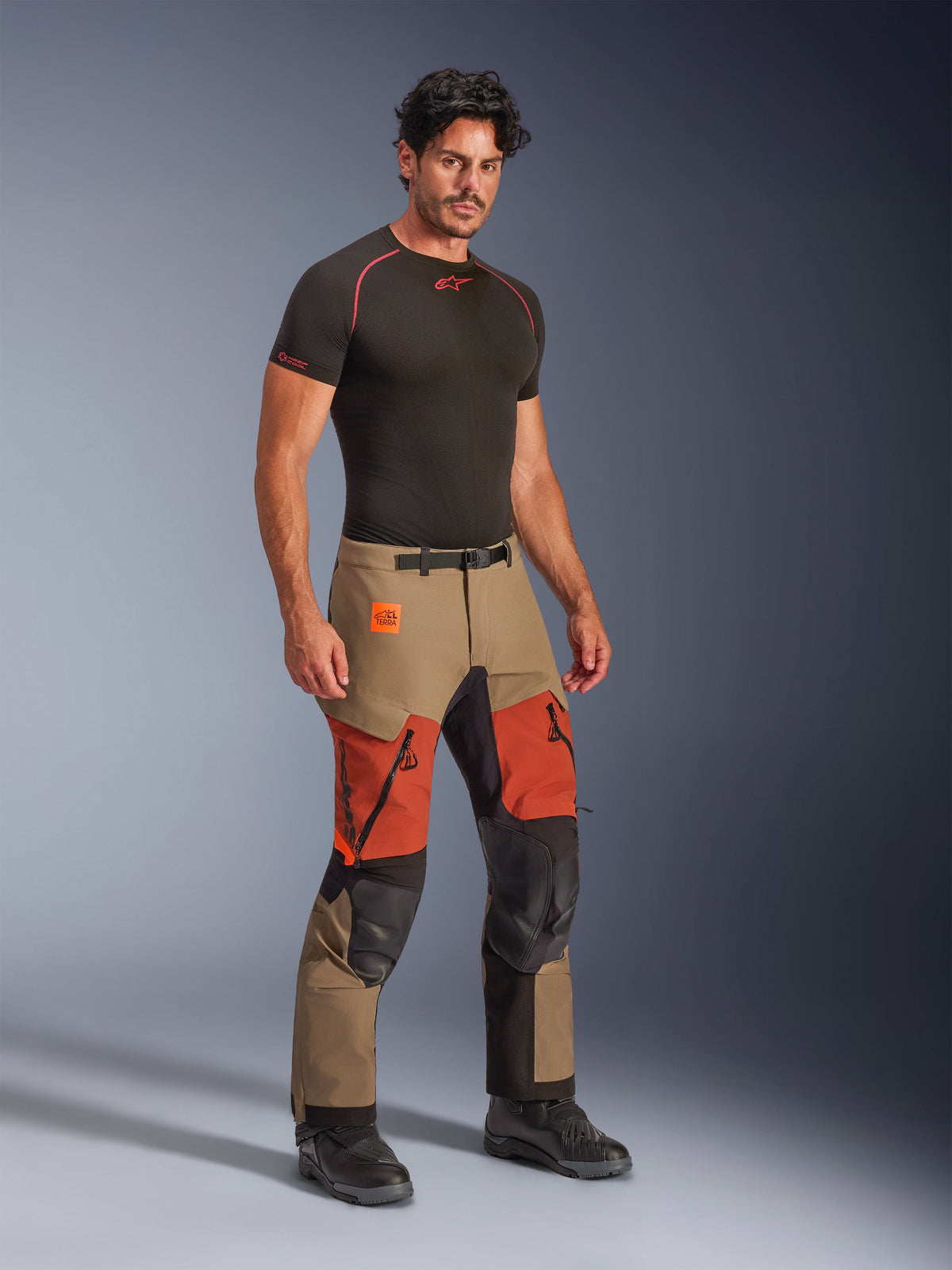 Alpinestars Halo Pro Drystar® XF Laminated Pants, Adventure Motorcycle Pants, Walnut Rust and Black, worn by a model with a black compression base layer and black boots, featuring reinforced knee panels, orange zippered thigh vents, and Terra branding, technical all-terrain riding gear