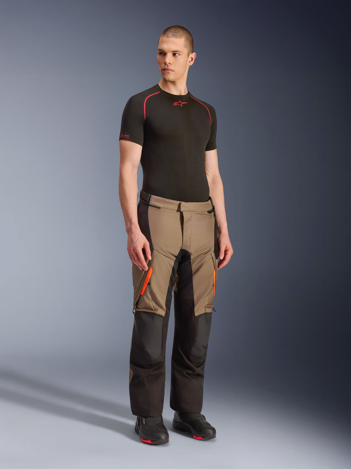 Alpinestars Andes V4 Drystar® bukser, adventure touring-bukser til motorcykel, Walnut Black med orange detaljer, vist på model med sort kortærmet baselayer og sorte støvler, med forstærkede tekstilpaneler og cargolommer, robust design til kørsel i al slags vejr