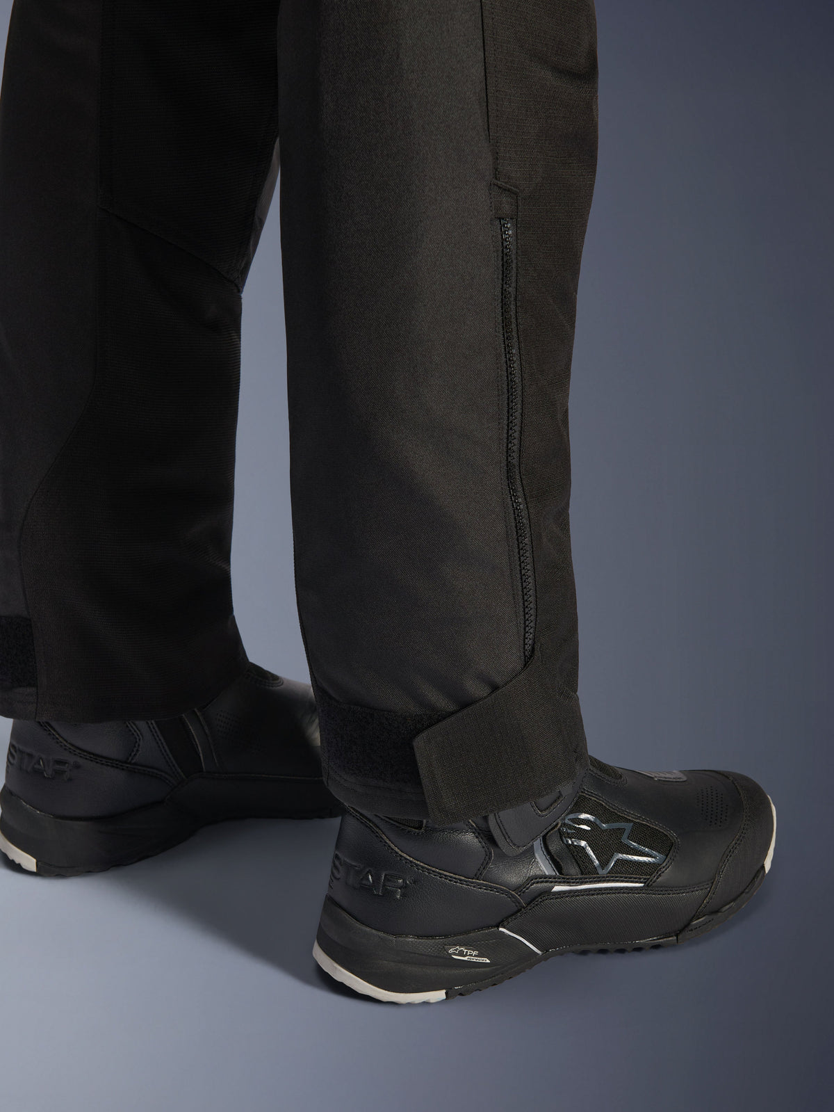 Alpinestars Stella Andes V4 Drystar® bukser, sort, nærbillede af underben og buksekant, med lynlås i siden og justerbar velcrostrop ved anklen, båret med sorte Alpinestars motorcykelstøvler
