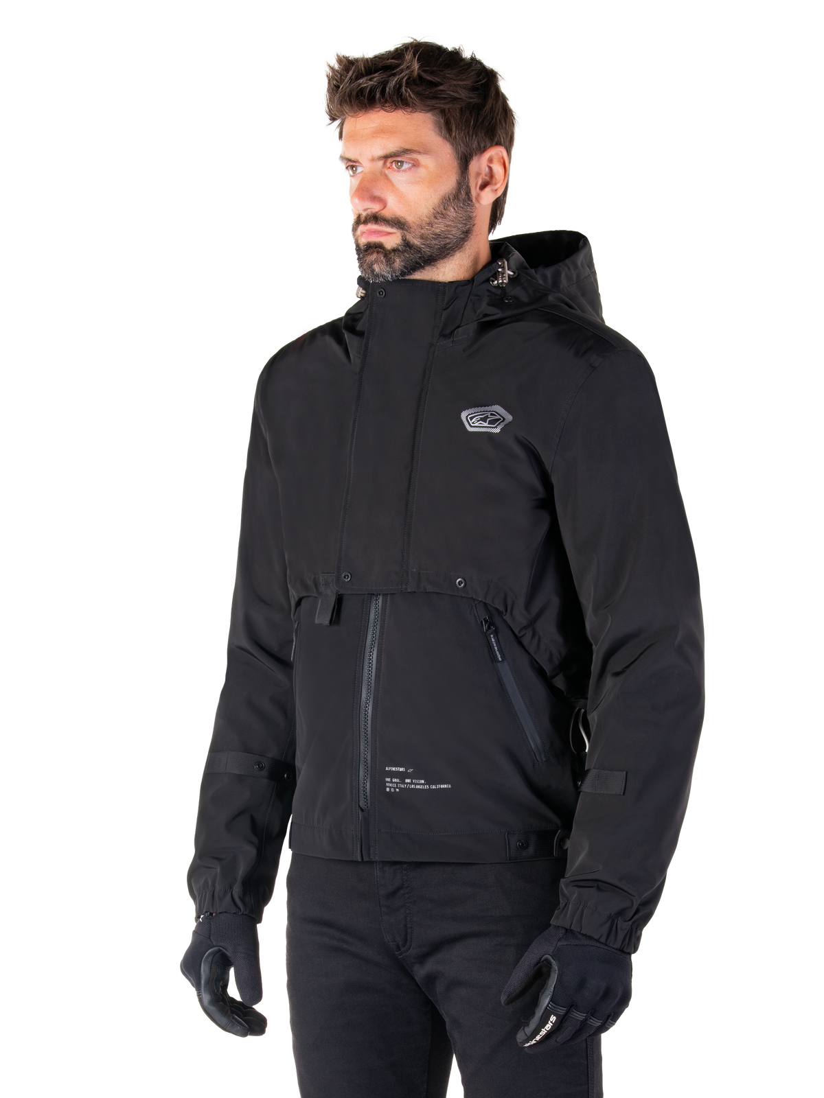 Alpinestars Mootant Vandtæt Parka, sort modulær motorcykeljakke, båret af en mand med sorte handsker og bukser, med hætte, lynlås foran og lagdelt utilitaristisk design, strømlinet urban æstetik med logo på brystet.