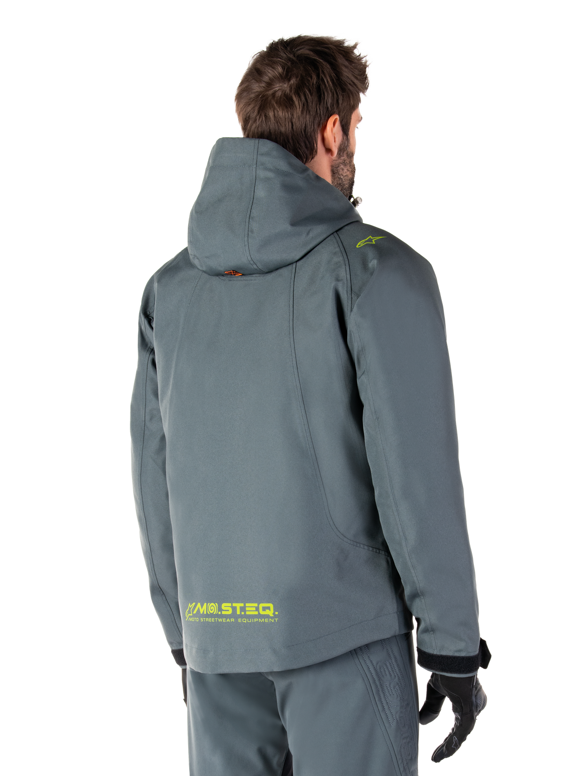 Alpinestars Morush Vandtæt Parka i Urban Gray, set bagfra, med gul MO.ST.EQ.-branding på lænden og skulderen, minimalistisk design med hætte, bæres med matchende grå kørebukser og sorte handsker, urban motorcykel-streetwear