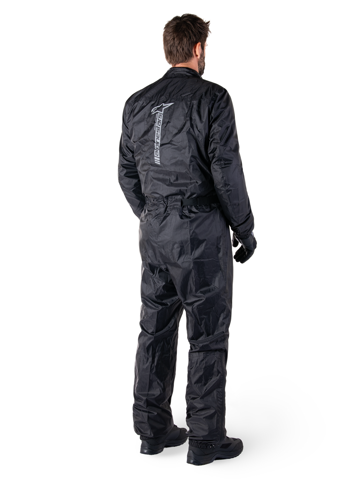 Alpinestars Hurricane Rain V2 Suit, sort vandtæt motorcykelregndragt i ét stykke, set bagfra med stort lodret reflekterende logo, letvægts vindafvisende stof, båret med sorte handsker og støvler