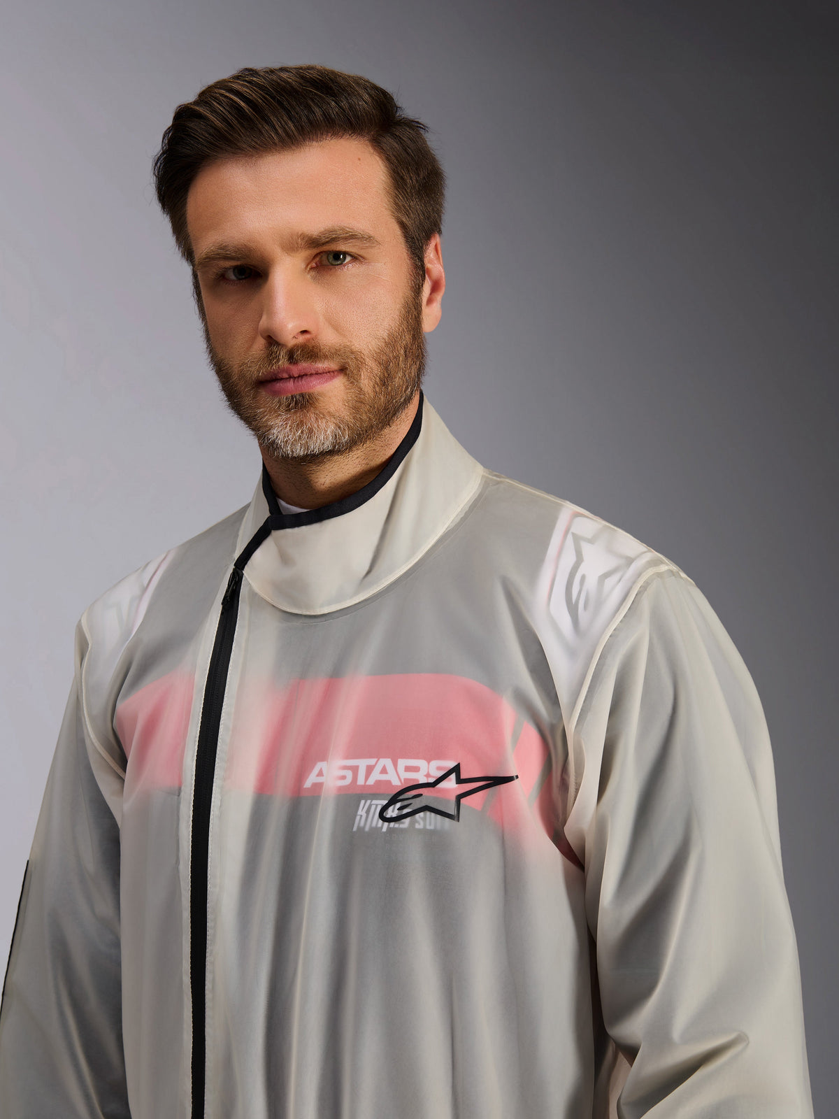 Alpinestars Kart Rain Suit, Karting Rain Oversuit, Clear, worn by a man, see-through waterproof material showing red and white racing suit details underneath, high collar with black zipper and Alpinestars star logo on the chest