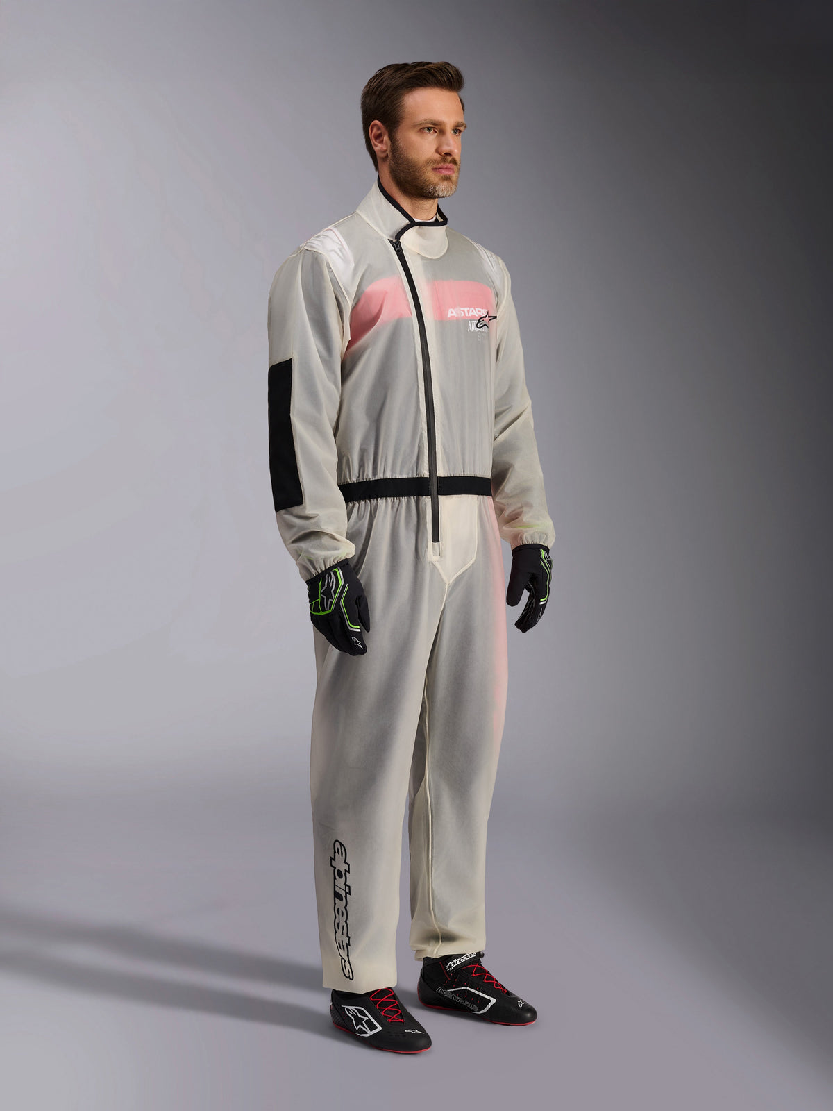 Alpinestars Kart Rain Suit, Clear waterproof karting oversuit, semi-transparent material showing a pink and white racing suit underneath, featuring a black asymmetric zipper, reinforced elbow patch, and black waist band, worn with black karting gloves and racing shoes.