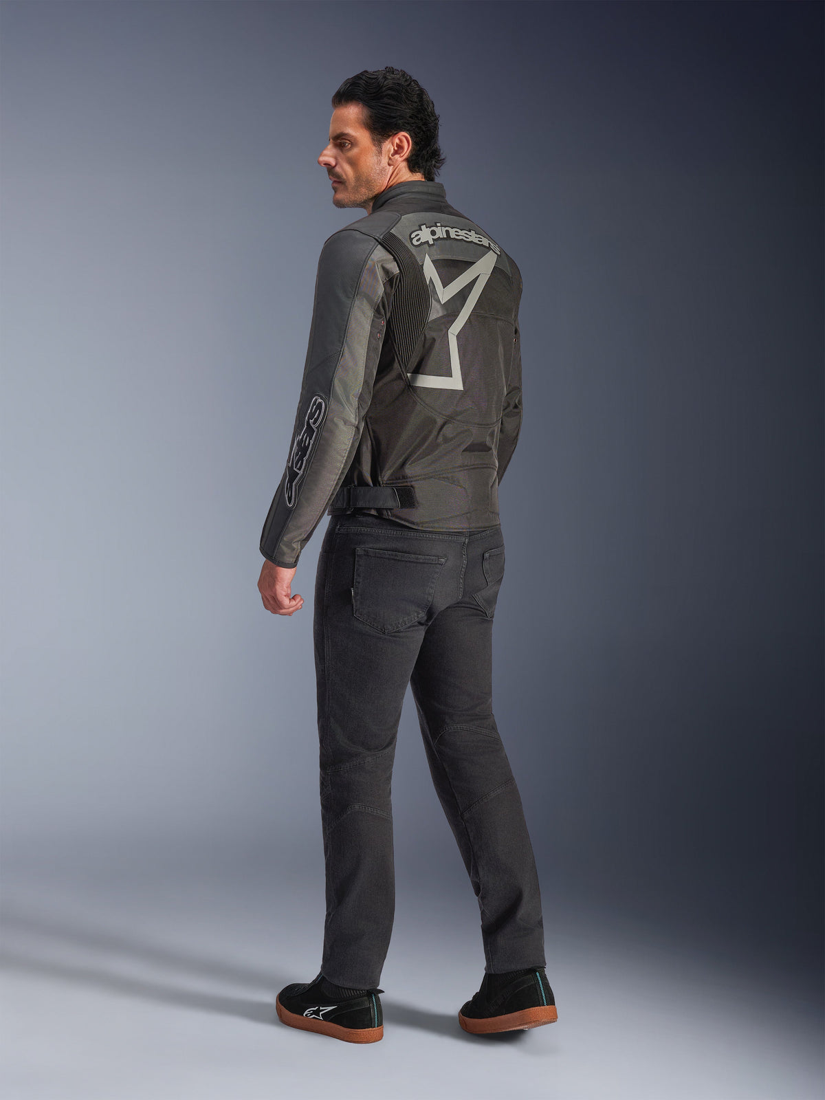 Alpinestars Axton Jacket, Men's Motorcycle Jacket, Black and Anthracite, back view featuring a large grey wrap-around logo and accordion shoulder panels, worn with dark riding pants and black sneakers, urban contemporary style with racing heritage.