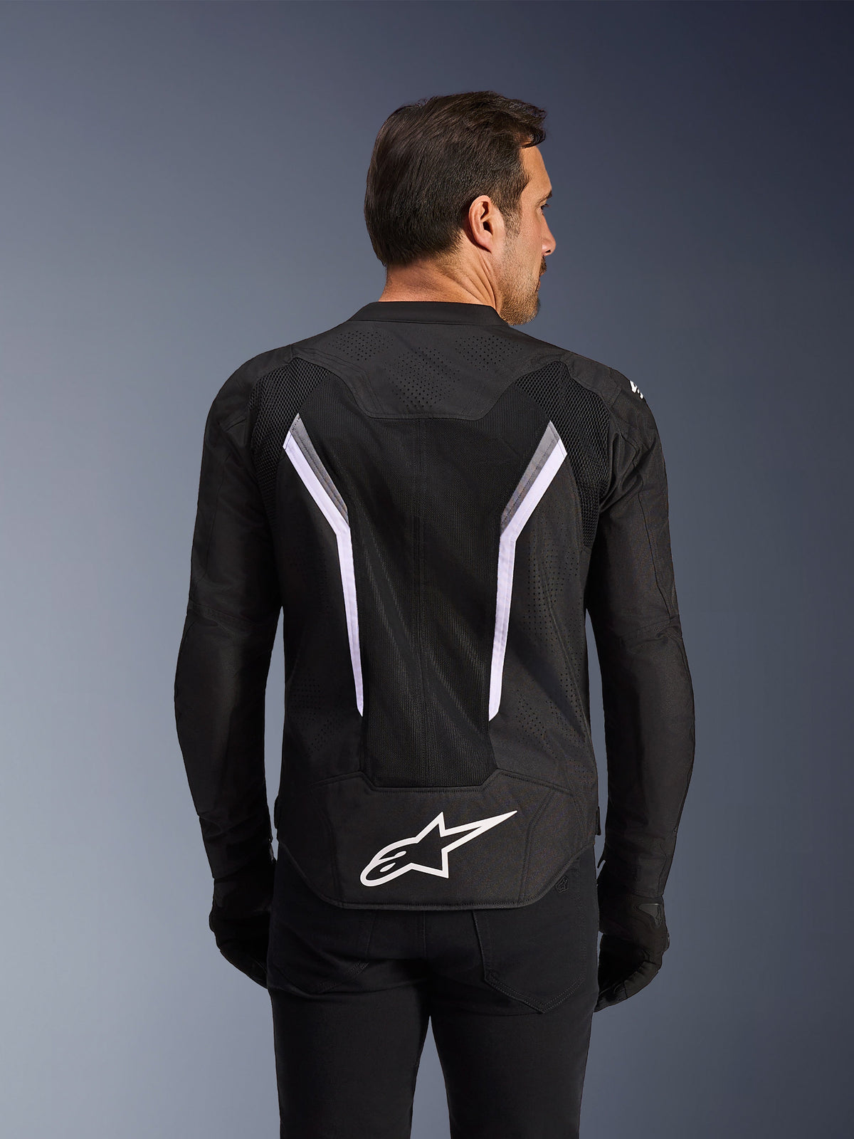 Alpinestars AST V3 Air Jakke, motorcykeljakke, sort med hvide og mørkegrå detaljer, set bagfra med omfattende mesh-ventilationspaneler, store hvide V-formede striber og hvidt Alpinestars-logo på den nederste del af ryggen, båret med sorte handsker og bukser