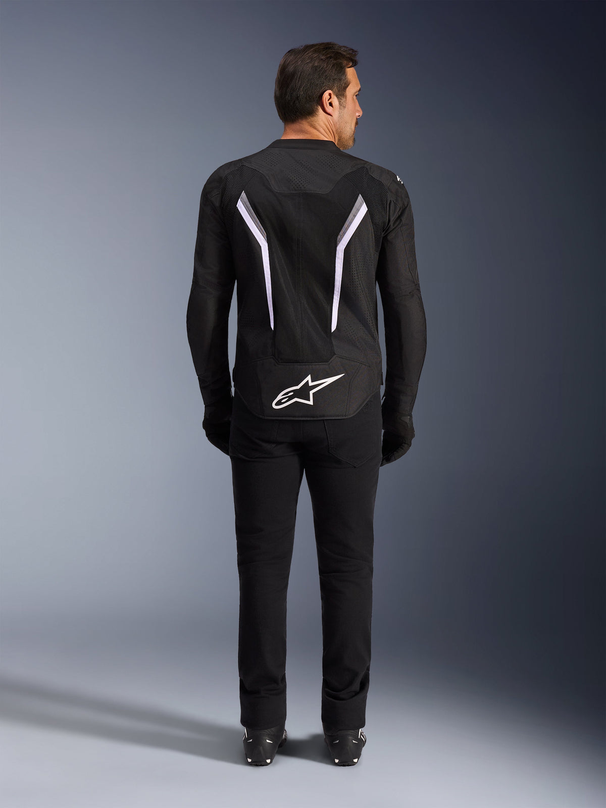 Alpinestars AST V3 Air-jakke, set bagfra, sort og mørkegrå med hvide detaljer, stort hvidt Astar-logo på lænden, refleksstriber på ryggen og mesh-tekstilkonstruktion, båret med sorte bukser og støvler for et sporty kørelook