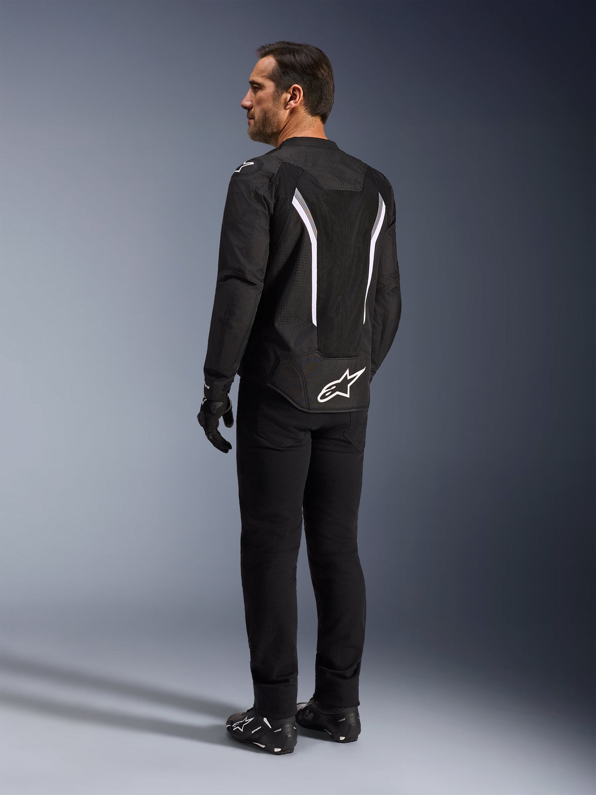 Alpinestars AST V3 Air-jakke, sort og hvid motorcykeljakke, set bagfra på model, med mesh-indsatser til luftgennemstrømning og hvidt logo nederst på ryggen, båret med sort teknisk køreudstyr