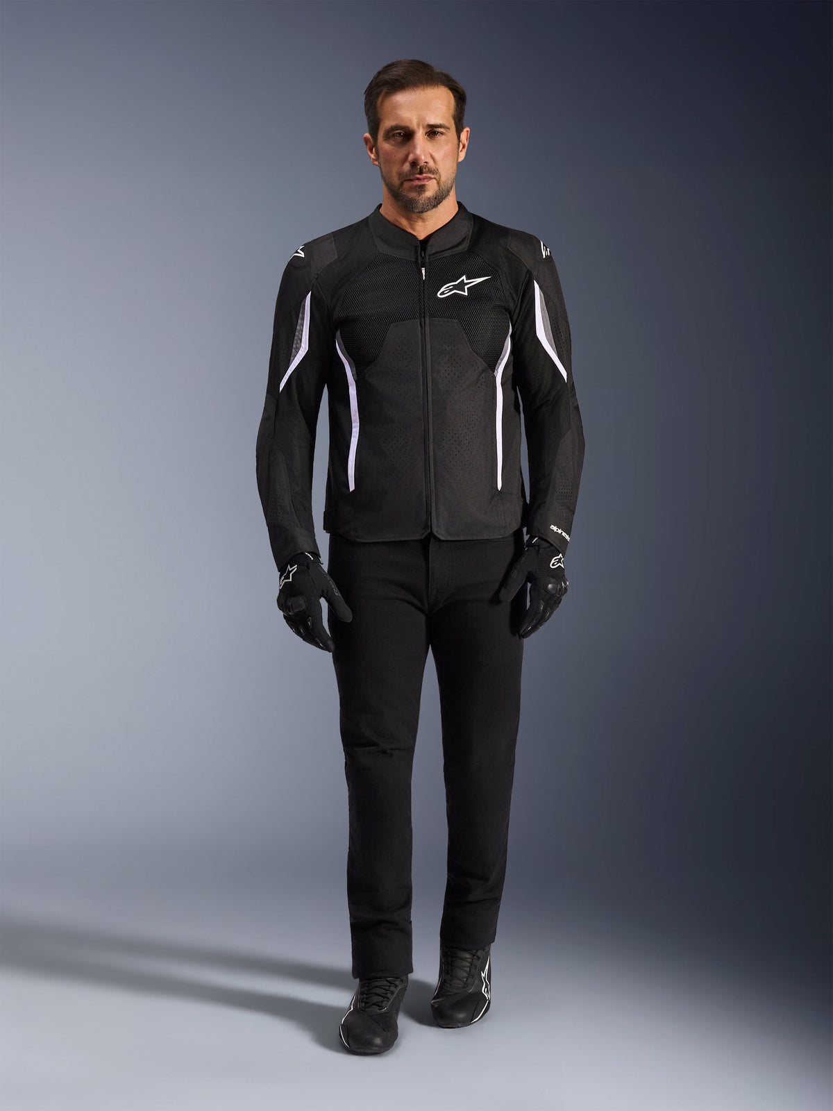Mandlig model iført Alpinestars AST V3 Air-jakke i sort med hvide detaljer, med ventilationspaneler i mesh og en sporty pasform, kombineret med sorte tekniske kørebukser, handsker og støvler, set forfra, teknisk motorcykeludstyr til kørsel i varmt vejr.