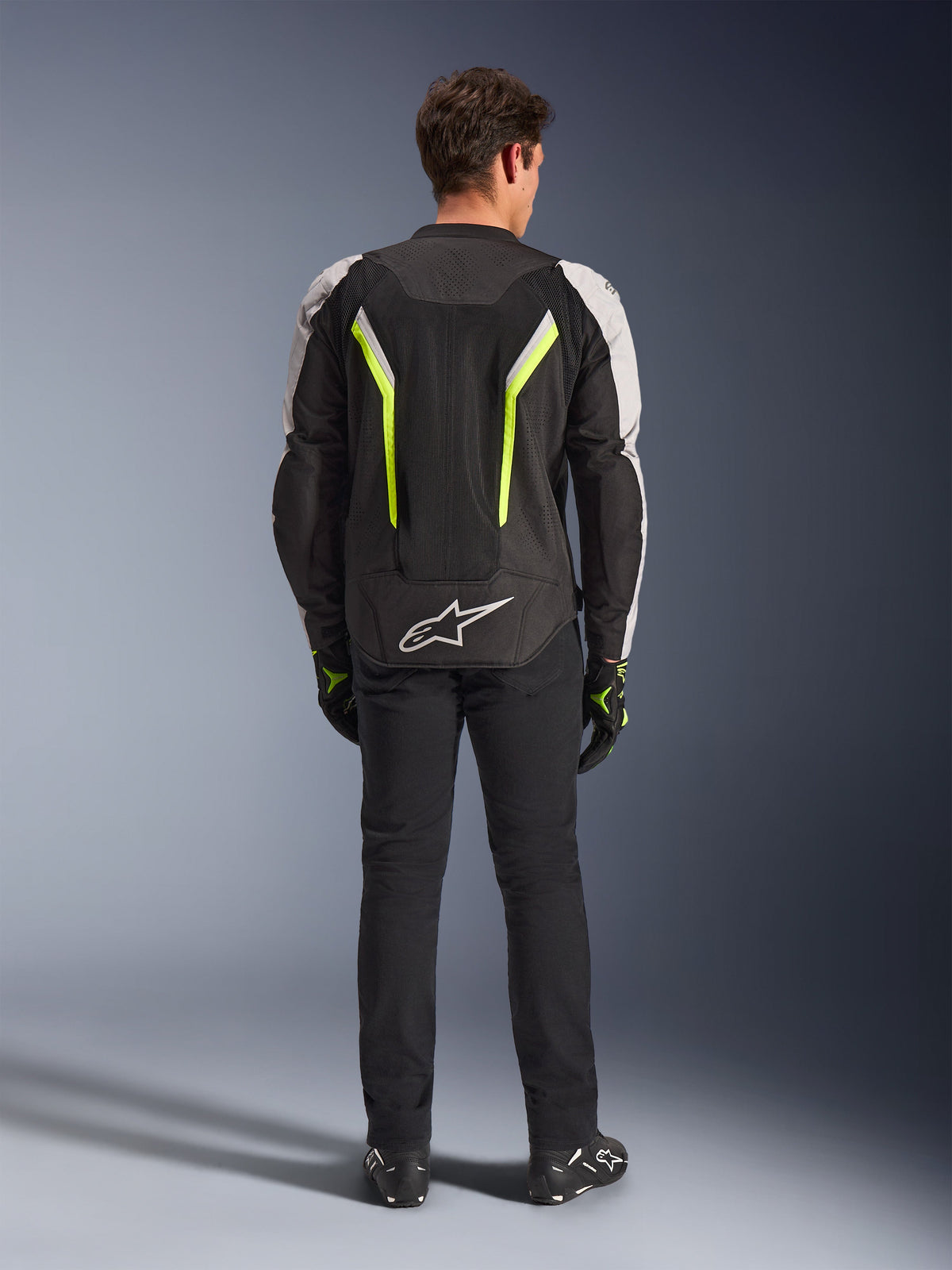Alpinestars AST V3 Air Jacket, tekstil motorcykeljakke, sort, sølv og gul fluo, set bagfra med mesh-ventilationspaneler og fluorescerende detaljer, båret med sorte bukser og kørehandsker, sportsligt køreudstyr