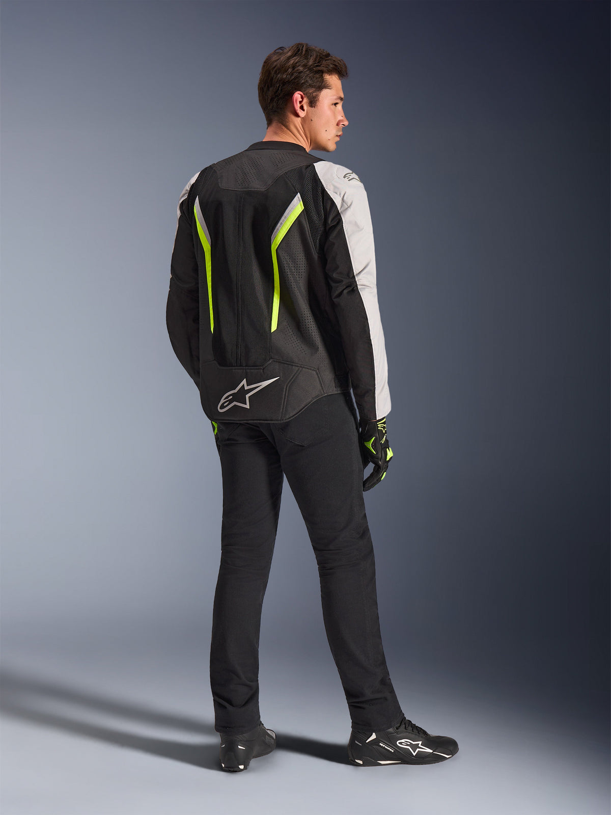 Alpinestars AST V3 Air-jakke i Black Silver Yellow Fluo, tekstil-motorcykeljakke båret af mandlig kører set bagfra, med omfattende mesh-paneler og fluorescerende gule detaljer, kombineret med sorte kørebukser, sorte handsker og sorte Alpinestars-sko, teknisk udstyr til sportskørsel.