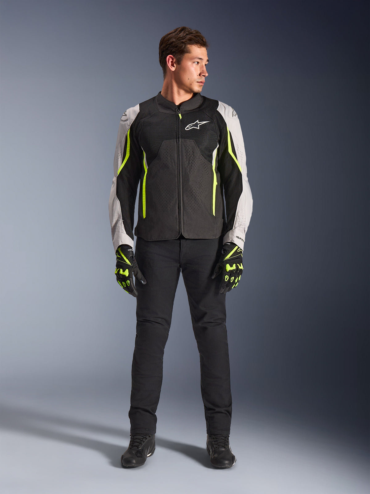 Mandlig model iført Alpinestars AST V3 Air-jakke i Black Silver Yellow Fluo, med mesh-ventilationspaneler og fluorescerende detaljer, kombineret med sorte kørebukser og matchende motorcykelhandsker, high-performance køreudstyr til sport.