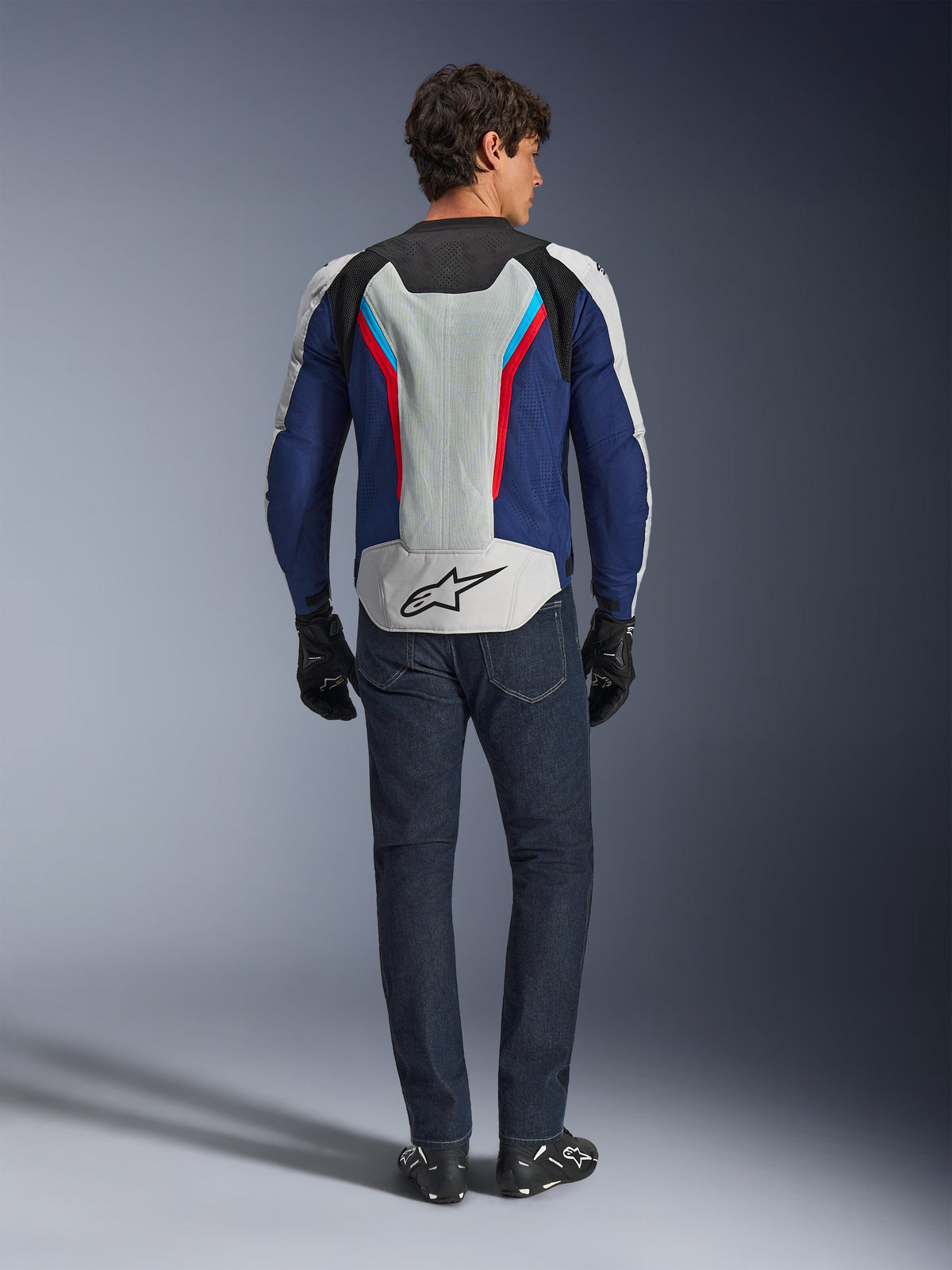 Alpinestars AST V3 Air-jakke, set bagfra, Silver Deep Blue og Bright Red, stort mesh-ventilationspanel på ryggen med Alpinestars-logo, båret med mørkeblå jeans og sorte handsker, sports-motorcykeludstyr til varmt vejr