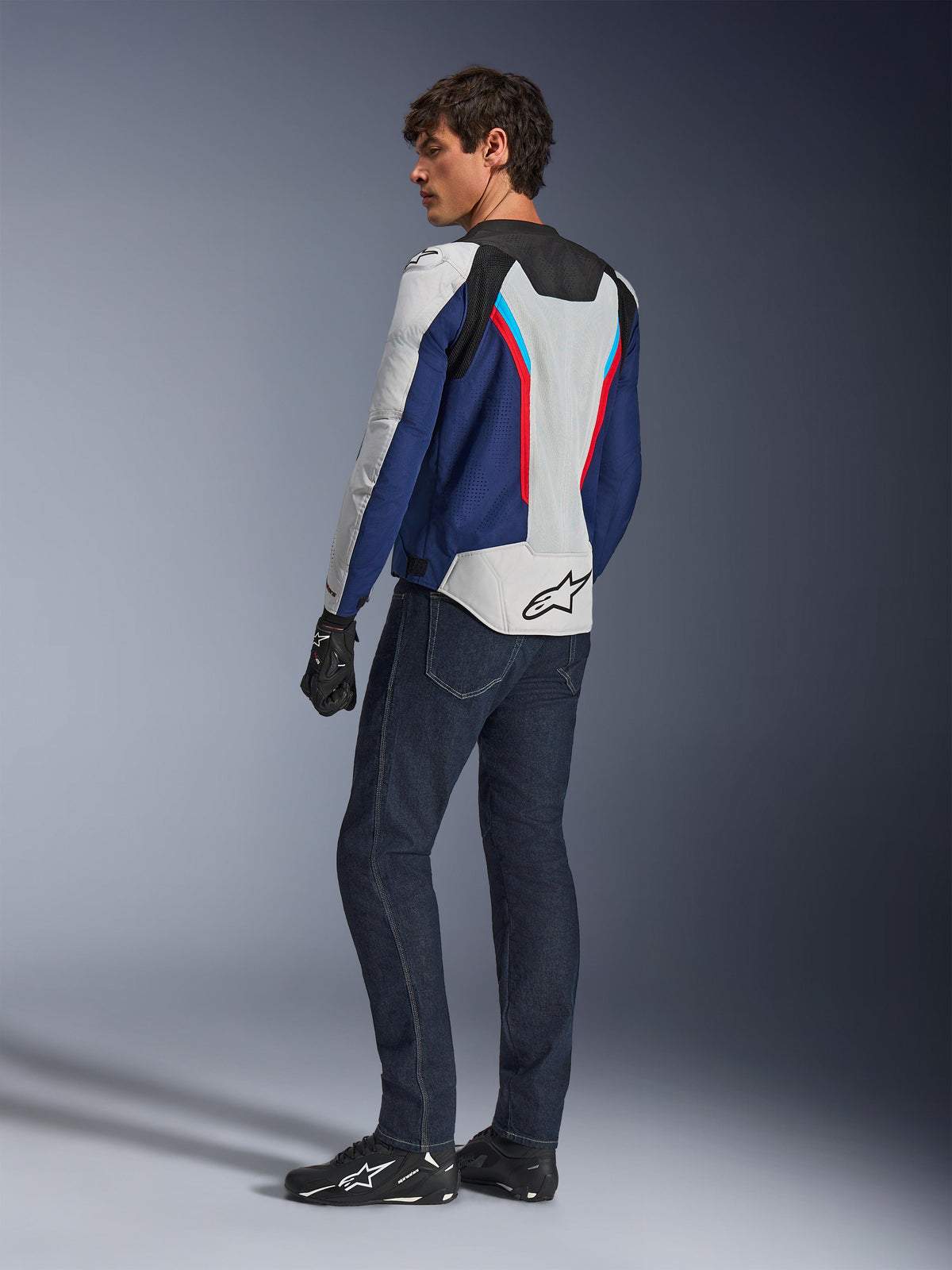 Alpinestars AST V3 Air-jakke, motorcykeljakke i Silver Deep Blue og Bright Red, set bagfra båret med mørkeblå kørejeans og sort udstyr, med åndbare mesh-paneler og Alpinestars-logo på ryggen, teknisk sport-fit køreudstyr.