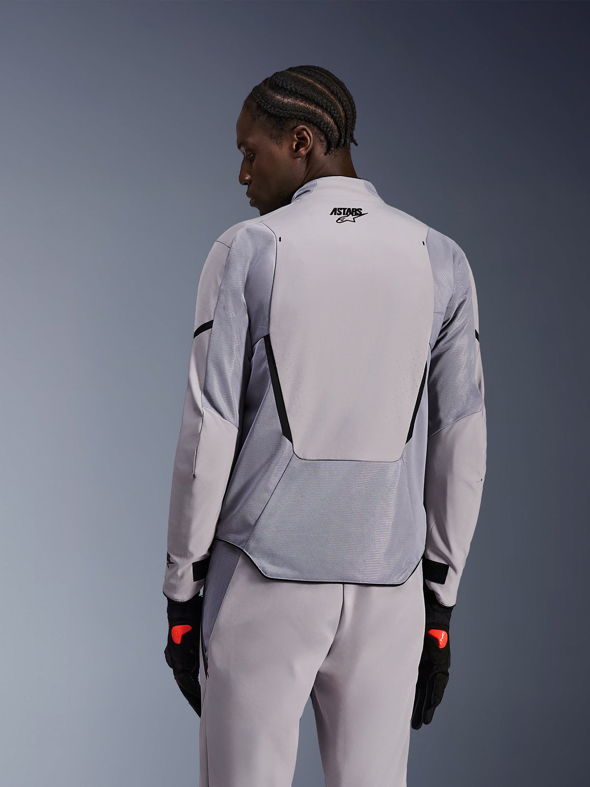 Alpinestars Aeroshell Airflow-jakke i Mid Gray Anthracite, set bagfra, åndbare mesh-paneler og laserperforeringer, teknisk stretch softshell-konstruktion med et forlænget rygpanel, båret med matchende grå bukser og sorte handsker for et sportsorienteret look