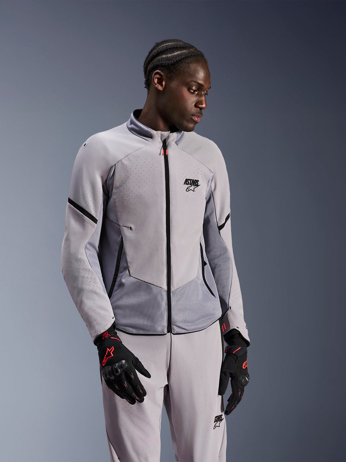 Alpinestars Aeroshell Airflow jakke, Mid Gray Anthracite, bæres af en model med matchende tekstilbukser og sorte handsker med røde detaljer, med laserperforerede zoner og mesh-paneler til ventilation, strømlinet sport-fit design til motorcykelkørsel.