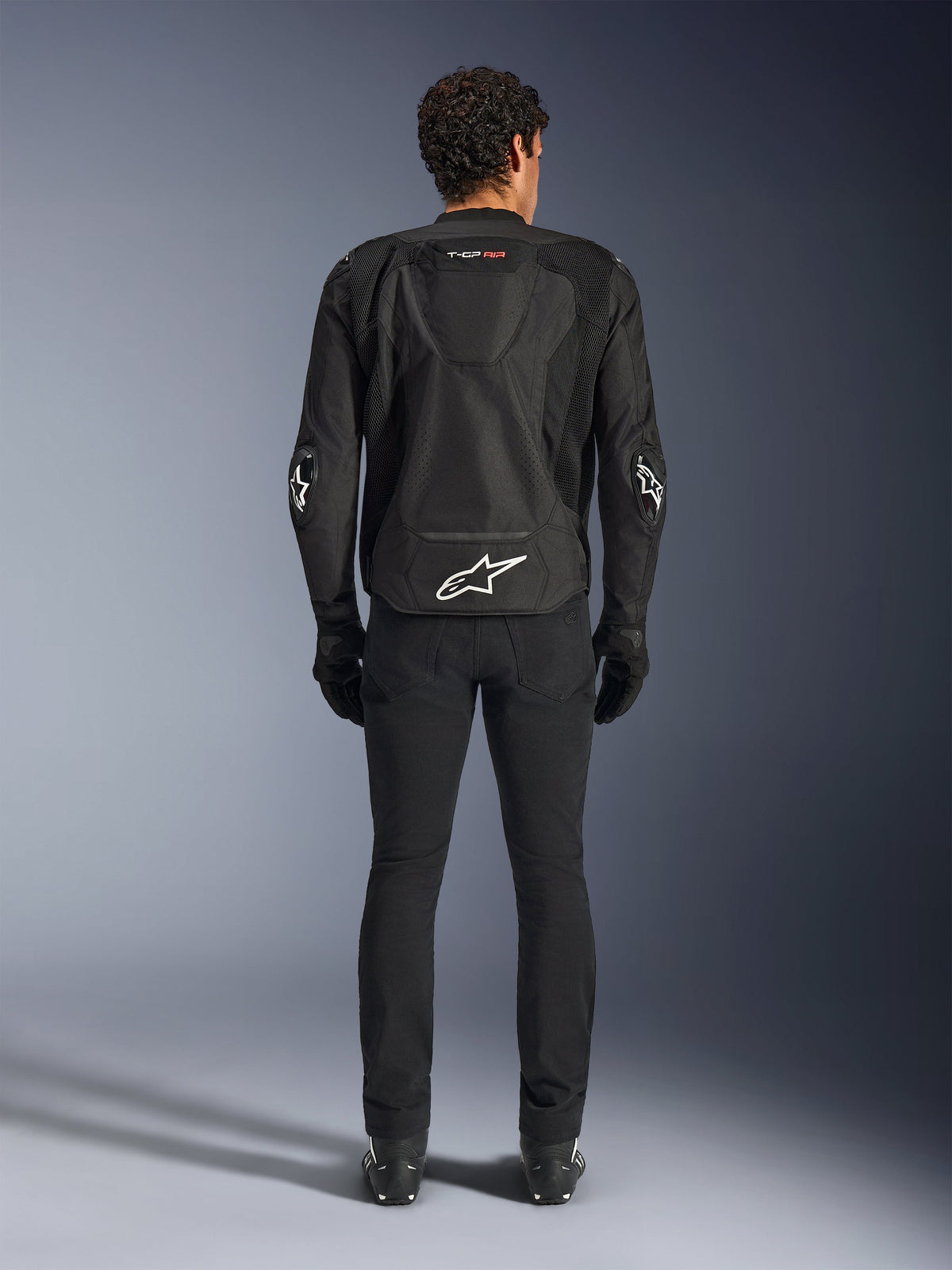 Alpinestars T-GP Air-jakke, sort, tekstil-motorcykeljakke set bagfra på en mand, med mesh-ventilationspaneler, en aerodynamisk pukkel og hvide Alpinestars-logoer på lænden og albuerne, båret med sorte motorcykelbukser og handsker