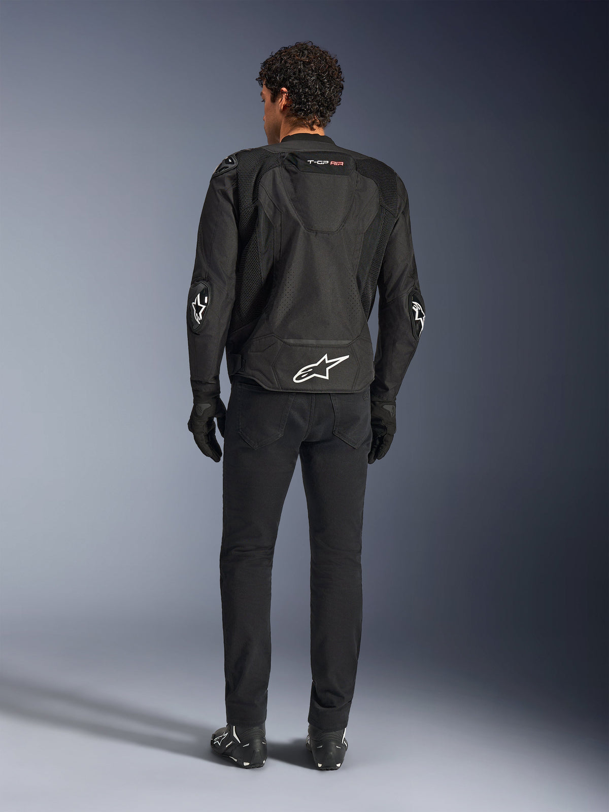 Alpinestars T-GP Air-jakke, sort, set bagfra på model, viser 3D-mesh ventilationspaneler og aerodynamisk pukkel, med hvidt stjernelogo på lænden og albuerne, båret med sorte motorcykelbukser og handsker, sportskøreudstyr.