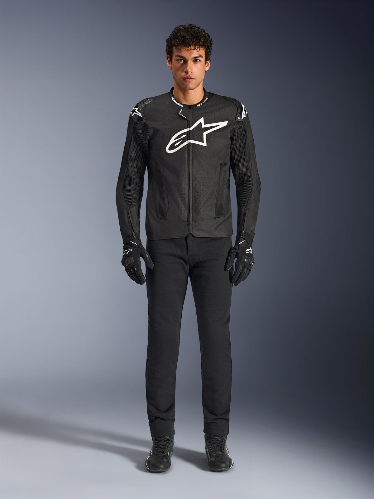 Alpinestars T-GP Air Jakke, sort tekstil motorcykeljakke med et stort hvidt Astars-logo på brystet, båret af en model med sorte kørebukser og handsker, med perforerede paneler og mesh-ventilation til sportskørsel