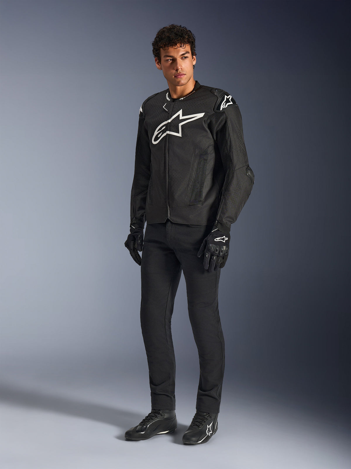 Alpinestars T-GP Air-jakke, sort tekstil-motorcykeljakke, båret af en model med matchende sorte bukser og handsker, med et fremtrædende hvidt Alpinestars-logo på brystet og skuldrene, ventilerede mesh-paneler og en sporty race-afledt pasform