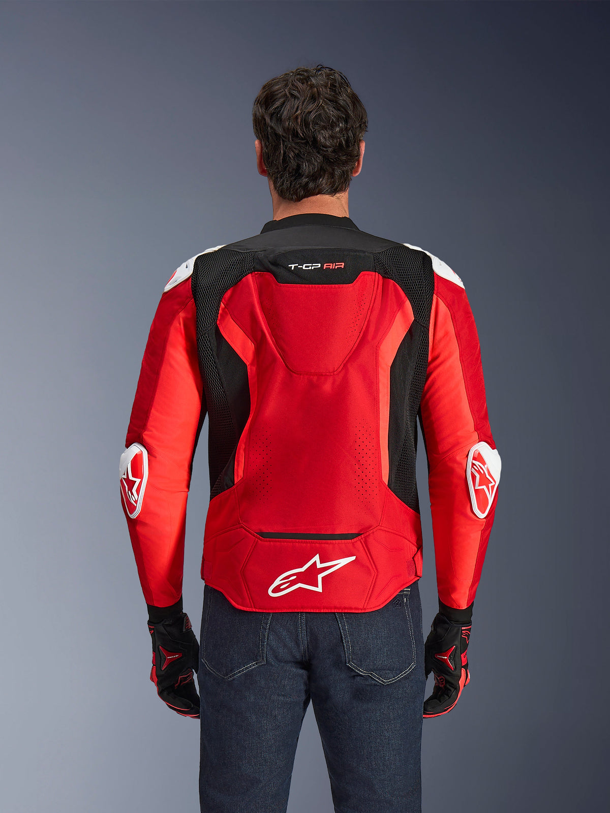 Alpinestars T-GP Air-jakke, tekstil-motorcykeljakke, klar rød og sort med hvide detaljer, set bagfra med den aerodynamiske pukkel og mesh-ventilationspaneler, Alpinestars-stjernelogo og T-GP Air-branding, båret med mørke jeans og racerhandsker.