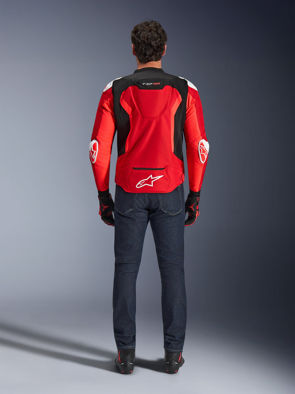 Alpinestars T-GP Air jakke, set bagfra, klar rød og sort, med hvidt logo nederst på ryggen og 'T-GP AIR' branding, båret med mørkeblå denimjeans og sorte og røde kørehandsker, sport-performance motorcykeludstyr