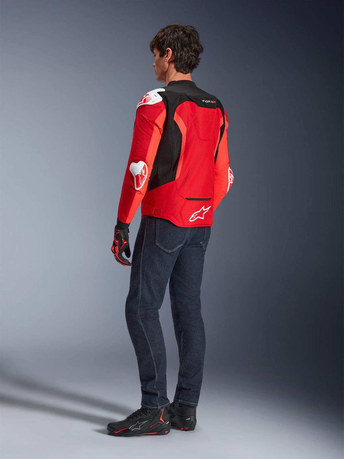 Alpinestars T-GP Air-jakke, set bagfra af en mand iført en stærk rød og fluorød tekstiljakke med sorte mesh-paneler og hvide beskyttelsesslidere, kombineret med mørkeblå jeans og sorte motorcykelsko, sportsmotorcykeludstyr