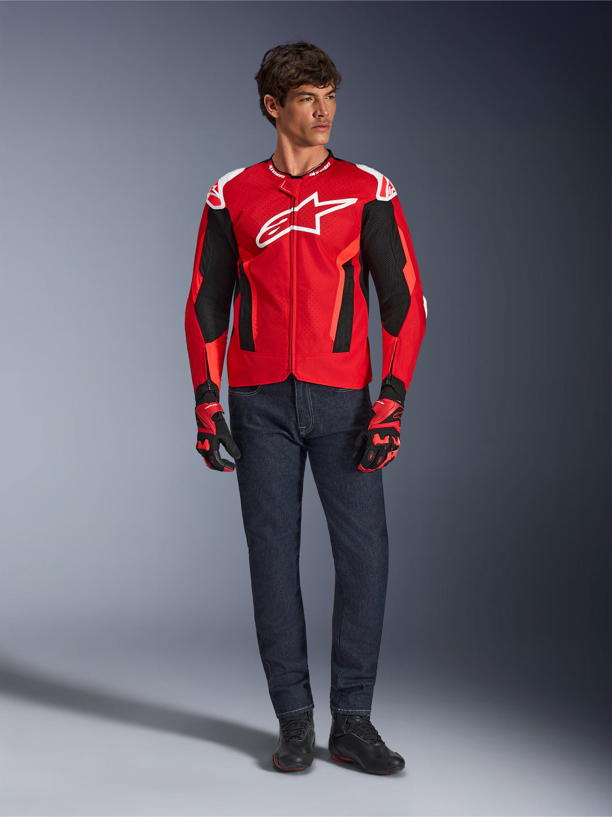 Alpinestars T-GP Air-jakke, Bright Red Red Fluo Black, mandlig model iført komplet motorcykeloutfit med mørkeblå jeans og matchende handsker, sports-tekstiljakke med perforerede mesh-paneler og hvidt Astars-logo, studiebillede.