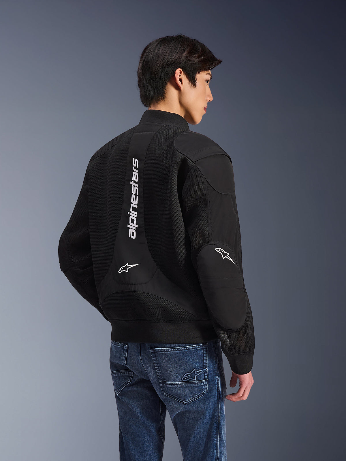 Alpinestars Flight Air Jacket, Black motorcycle jacket, back view showing large vertical white logo and mesh ventilation panels, bomber-style with ribbed hem, worn with blue jeans, urban contemporary riding gear