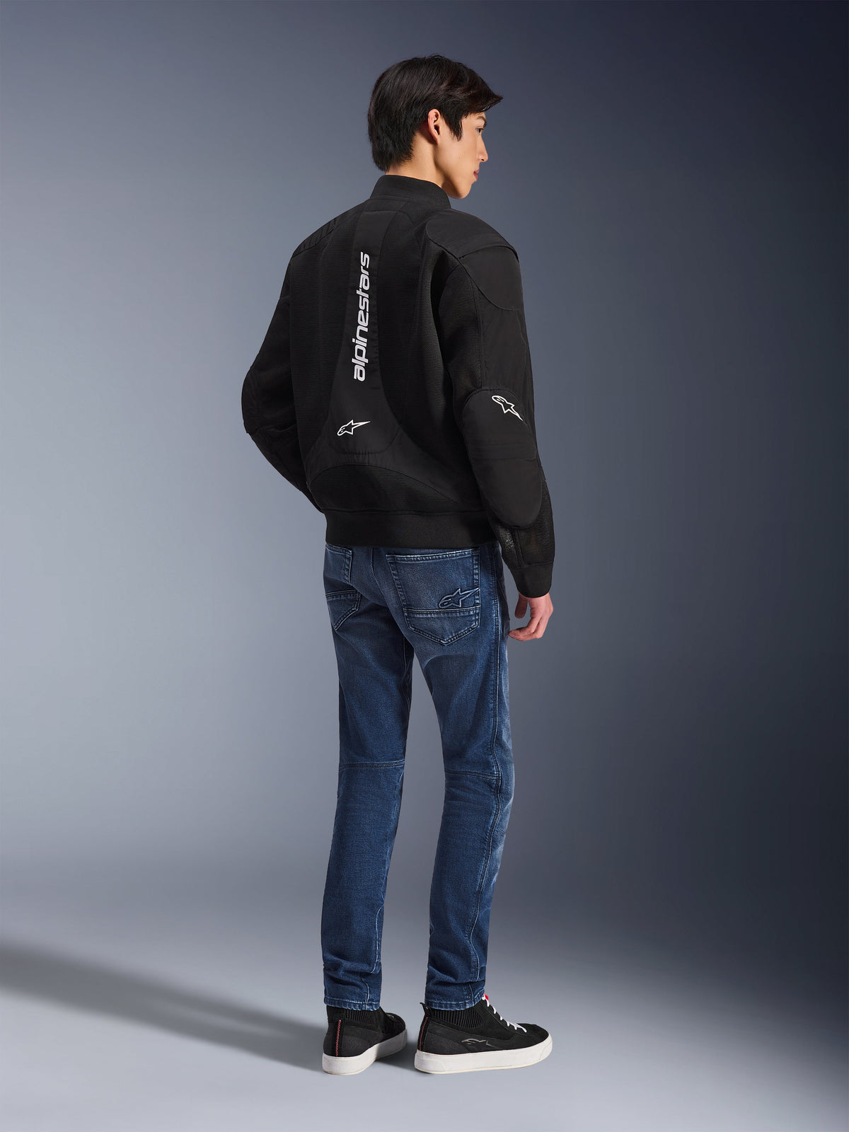 Model wearing Alpinestars Flight Air Jacket in Black, viewed from the back, paired with blue riding jeans and black sneakers, featuring vertical white Alpinestars branding and 3D mesh ventilation panels.