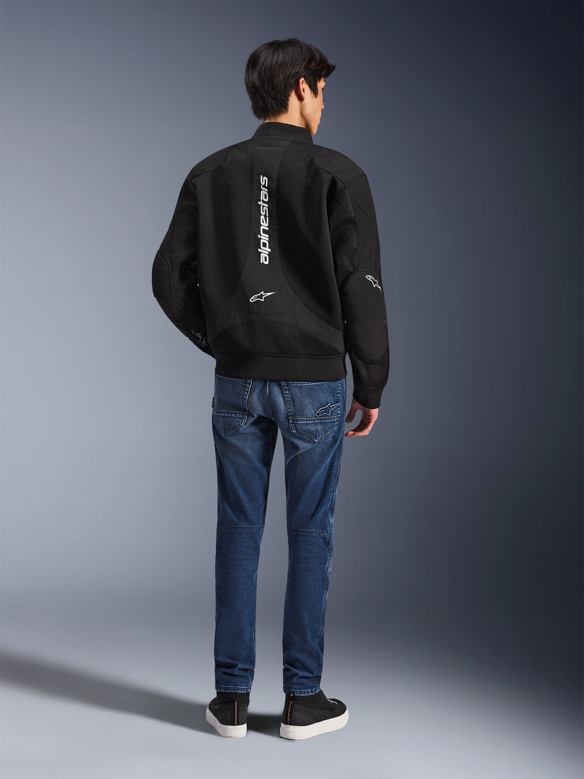 Model wearing the Alpinestars Flight Air Jacket from the back, Black bomber-style motorcycle jacket with 3D mesh panels and vertical white logo, paired with blue denim riding jeans and black sneakers, contemporary urban motorcycle gear