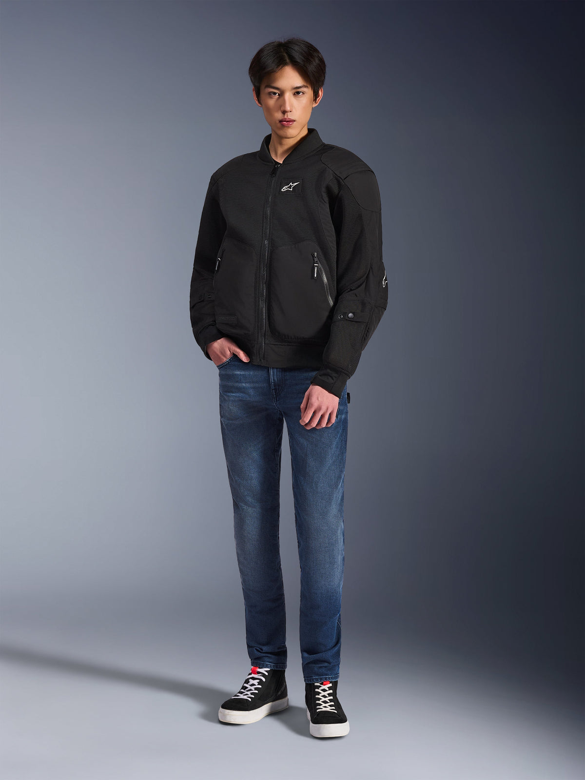 Model wearing the Alpinestars Flight Air Jacket in Black, an urban bomber-style motorcycle jacket with 3D mesh panels and a front zipper, paired with blue jeans and black sneakers, contemporary street-inspired riding apparel.