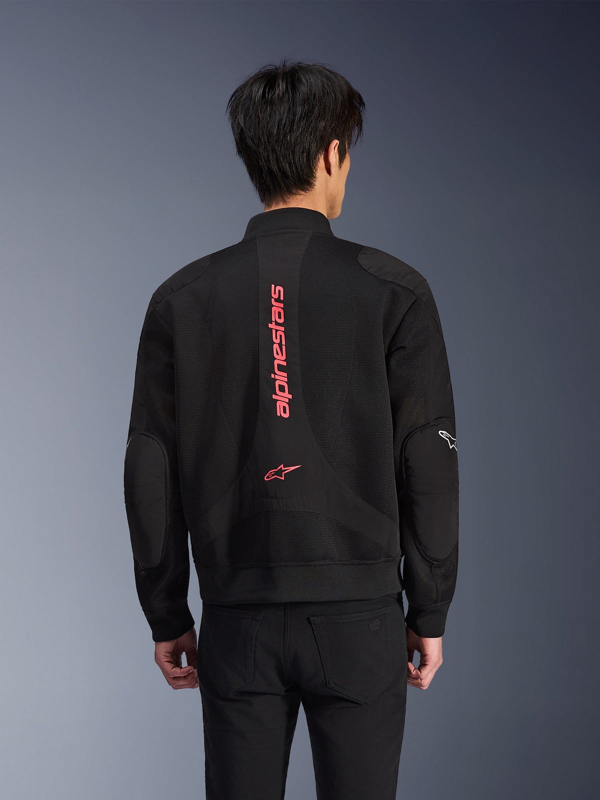 Set bagfra af en kører iført Alpinestars Flight Air-jakke i Black Bright Red, urban motorcykeljakke i bomber-stil med 3D-mesh-konstruktion, med stor lodret rød Alpinestars-branding og stjerne-logo på ryggen, gadeinspireret teknisk udstyr