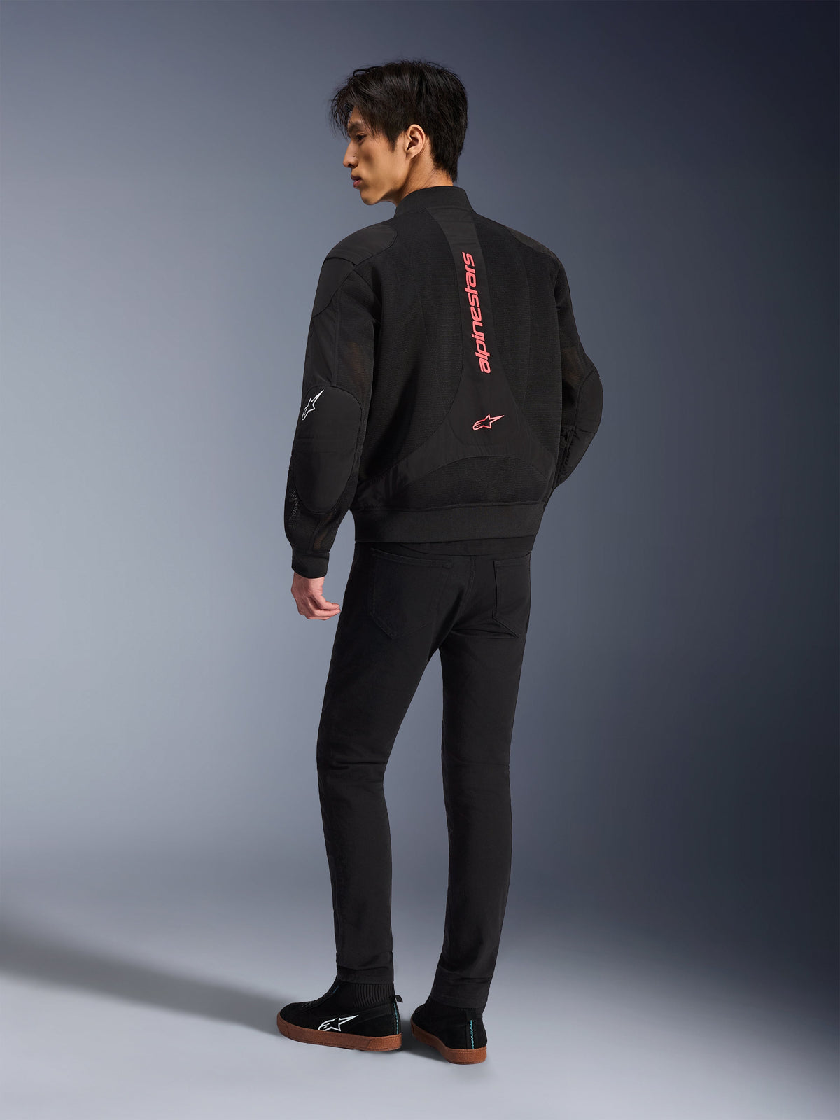 Mandlig model iført Alpinestars Flight Air-jakke i Black Bright Red, set bagfra viser lodret rød branding på ryggen og 3D-mesh-konstruktion, kombineret med sorte bukser og sneakers, urban motorcykelstil
