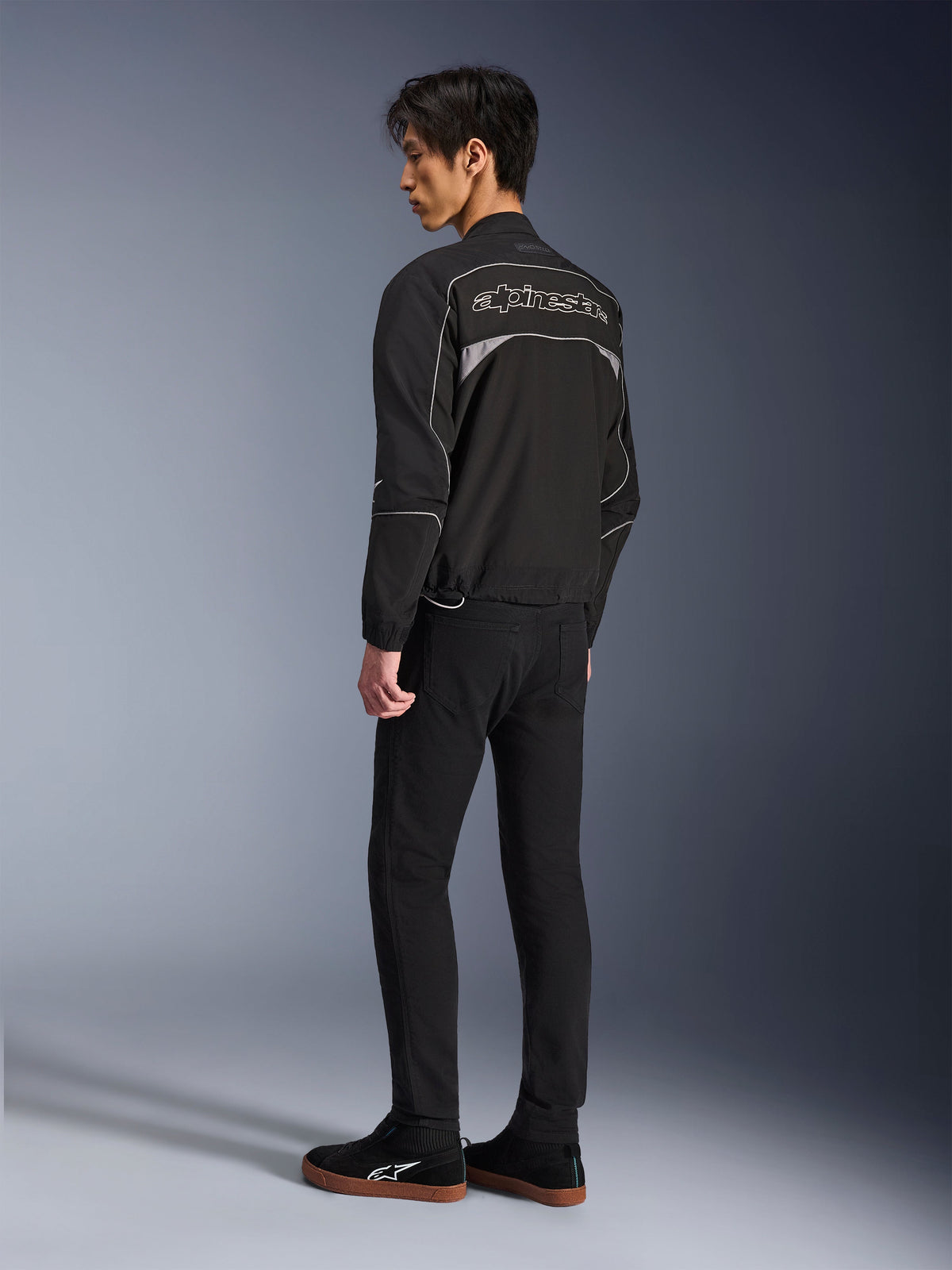Alpinestars Unite Jacket, Urban Motorcycle Jacket, Black and Dark Gray, back view showing large outlined Alpinestars logo, reflective piping and panels, minimalist softshell design, worn with black pants and casual footwear