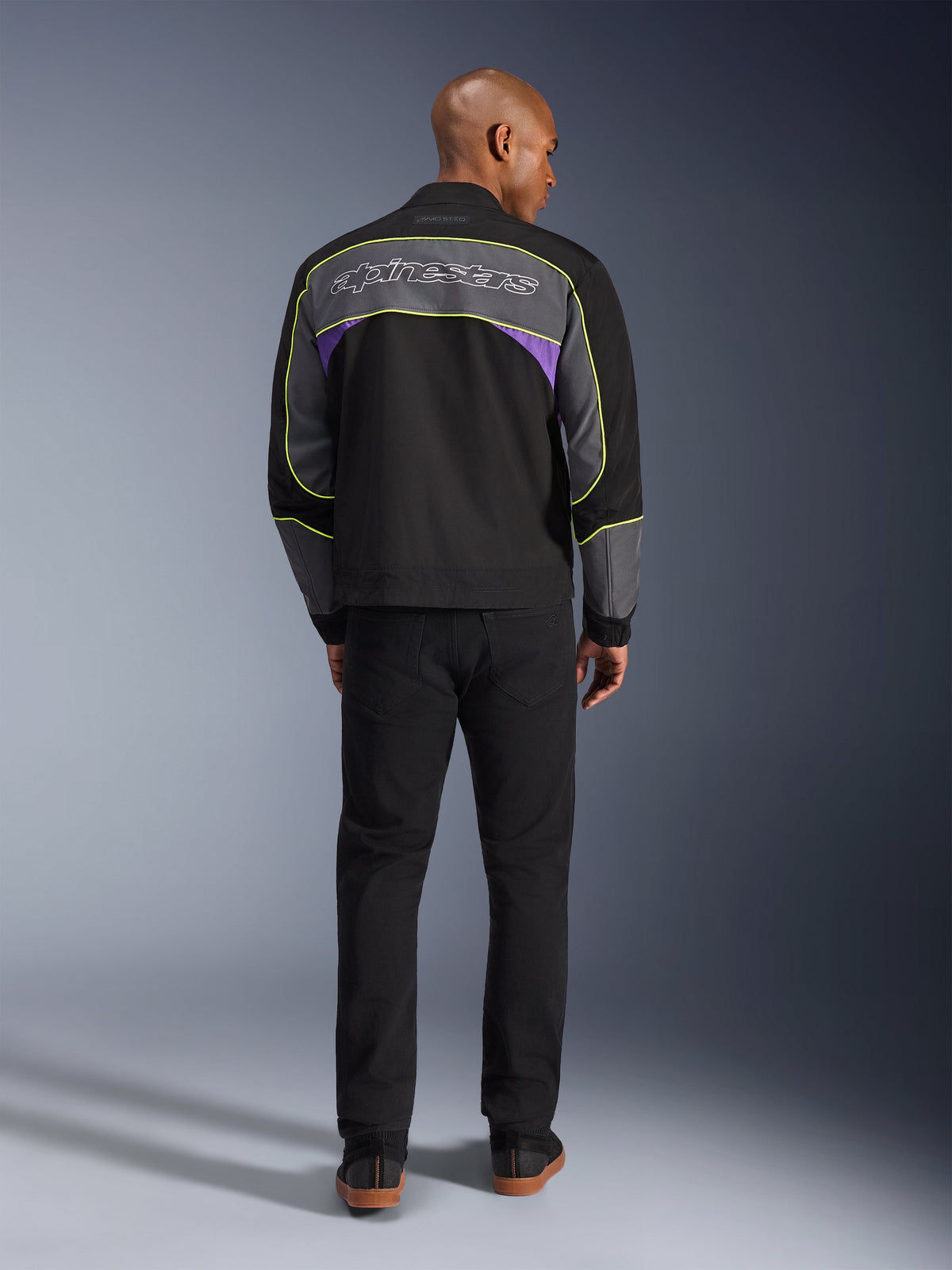 Alpinestars Unite-jakke, urban motorcykeljakke i Black Asphalt Violet, set bagfra med grå rygpaneler, lilla accenter og neongul piping, Alpinestars-logo hen over skuldrene, båret med sorte bukser