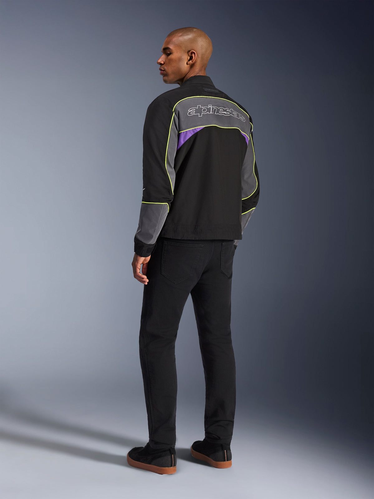 Alpinestars Unite Jakke, urban motorcykeljakke, Black Asphalt Violet, trefjerdedels visning bagfra på model, sort softshell med grå paneler og lilla accenter, neongule reflekterende kanter, Alpinestars-logo på den øverste del af ryggen, kombineret med sorte bukser og sorte fritidssko