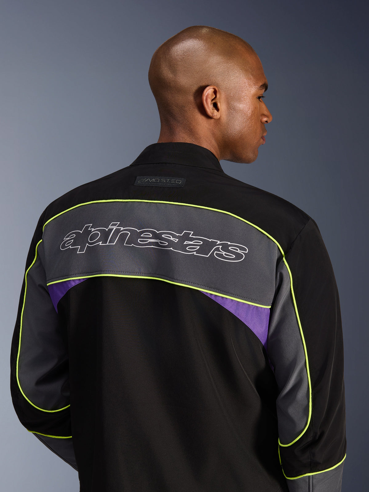 Alpinestars Unite jakke, urban motorcykeljakke, Black Asphalt Violet, set bagfra viser et stort gråt skulderpanel med Alpinestars-logo, neongul piping og lilla detaljer på en 3-lags softshell-konstruktion