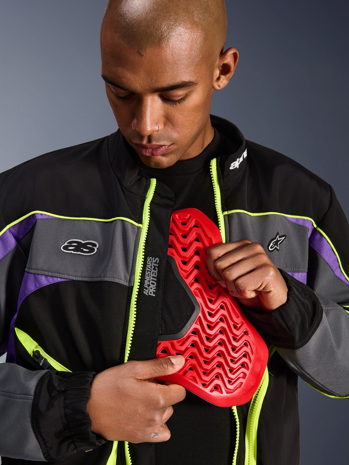 Alpinestars Unite-jakke, urban motorcykeljakke, Black Asphalt og Violet med neongule detaljer, mandlig model der viser en rød Nucleon brystpanser-indsats inde i den åbne jakke, minimalistisk streetwear-æstetik til bykørsel.