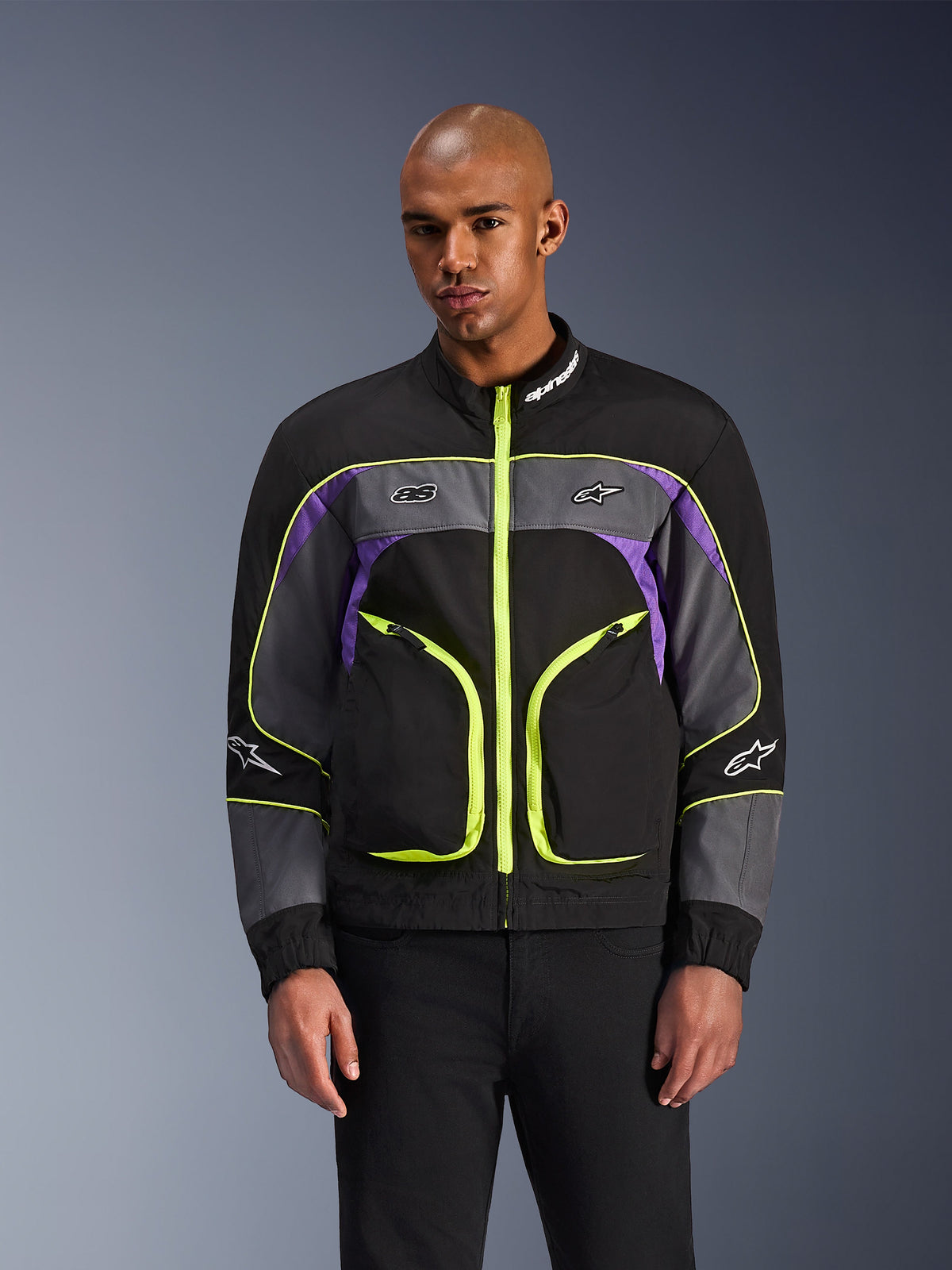 Alpinestars Unite jakke, urban motorcykeljakke, Black Asphalt og Violet med neongule detaljer, store frontlommer med neongule lynlåse, høj krave med hvidt logo, vist på model, moderne streetwear-inspireret beskyttelsesudstyr