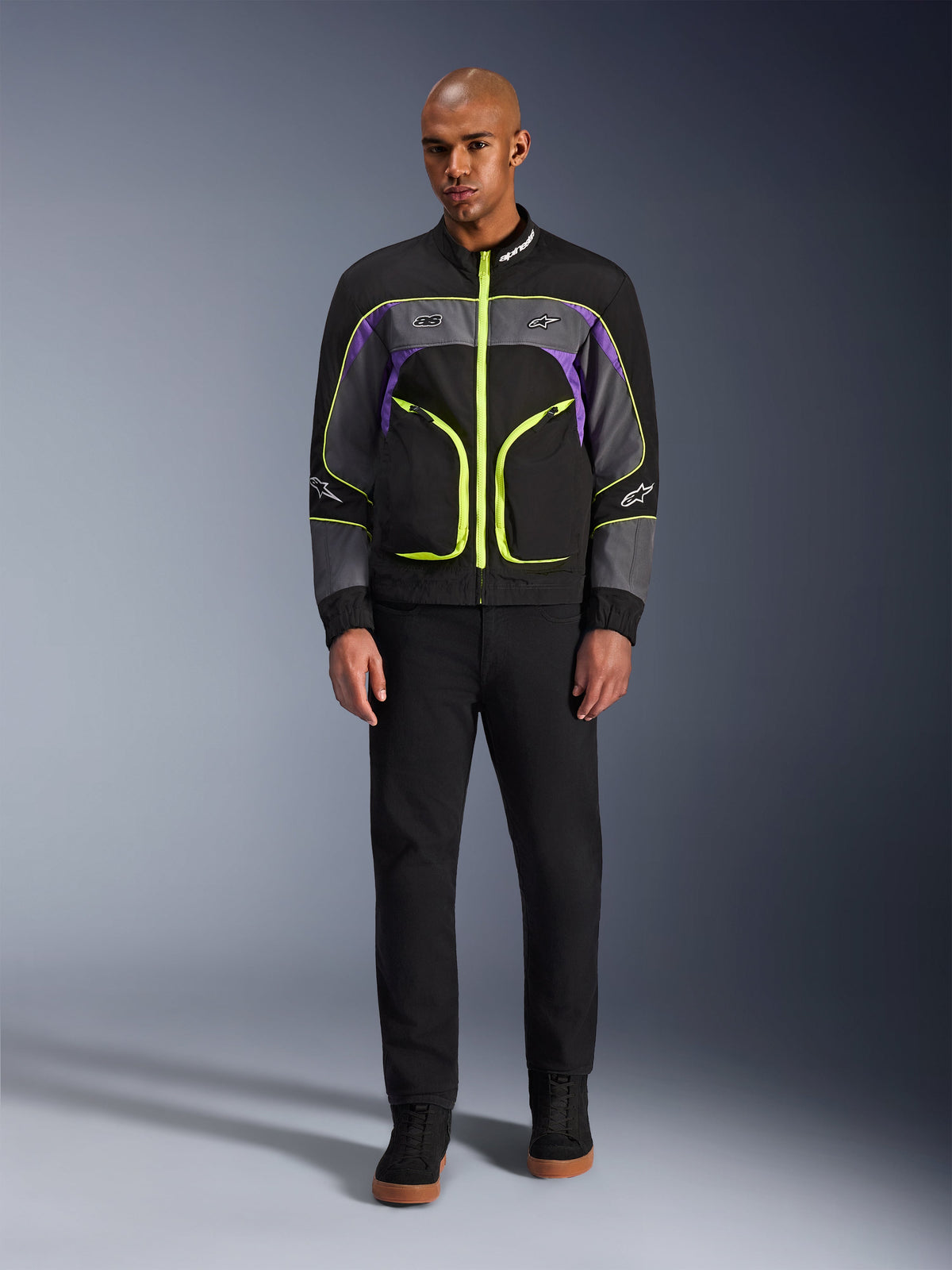 Mandlig model iført Alpinestars Unite-jakken i Black Asphalt Violet, med sort base, grå og lilla paneler og iøjnefaldende neongule lynlåse og detaljer, kombineret med sorte bukser og sko, minimalistisk urban motorcykeljakke.