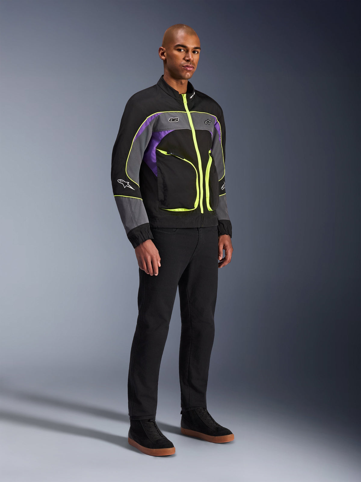 Alpinestars Unite jakke, urban motorcykeljakke, Black Asphalt Violet med neongule detaljer, båret af model med sorte bukser og sneakers, moderne streetwear-stil til bykørsel