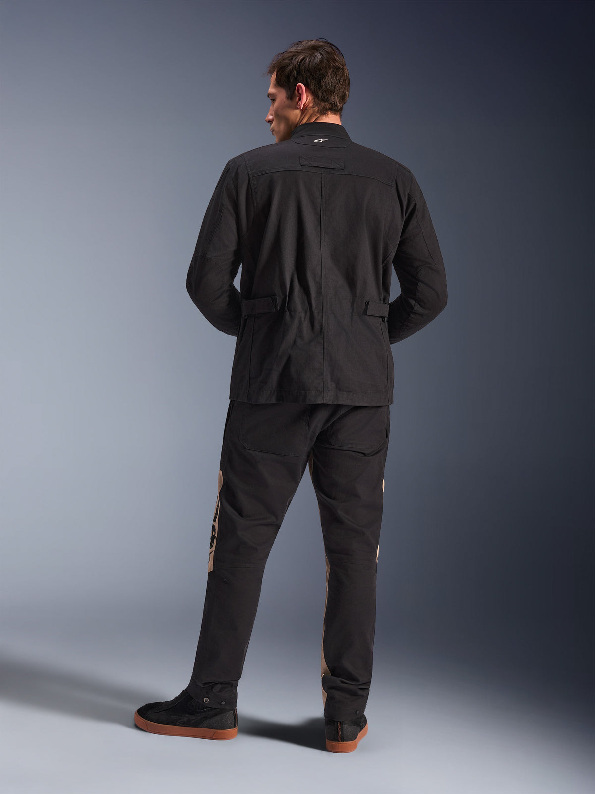 Alpinestars Dice-jakke set bagfra, sort, urban motorcykel-feltjakke med plisserede skuldre og taljejusteringer, parret med sorte og sandfarvede kørebukser og sneakers, moderne stil til bypendling