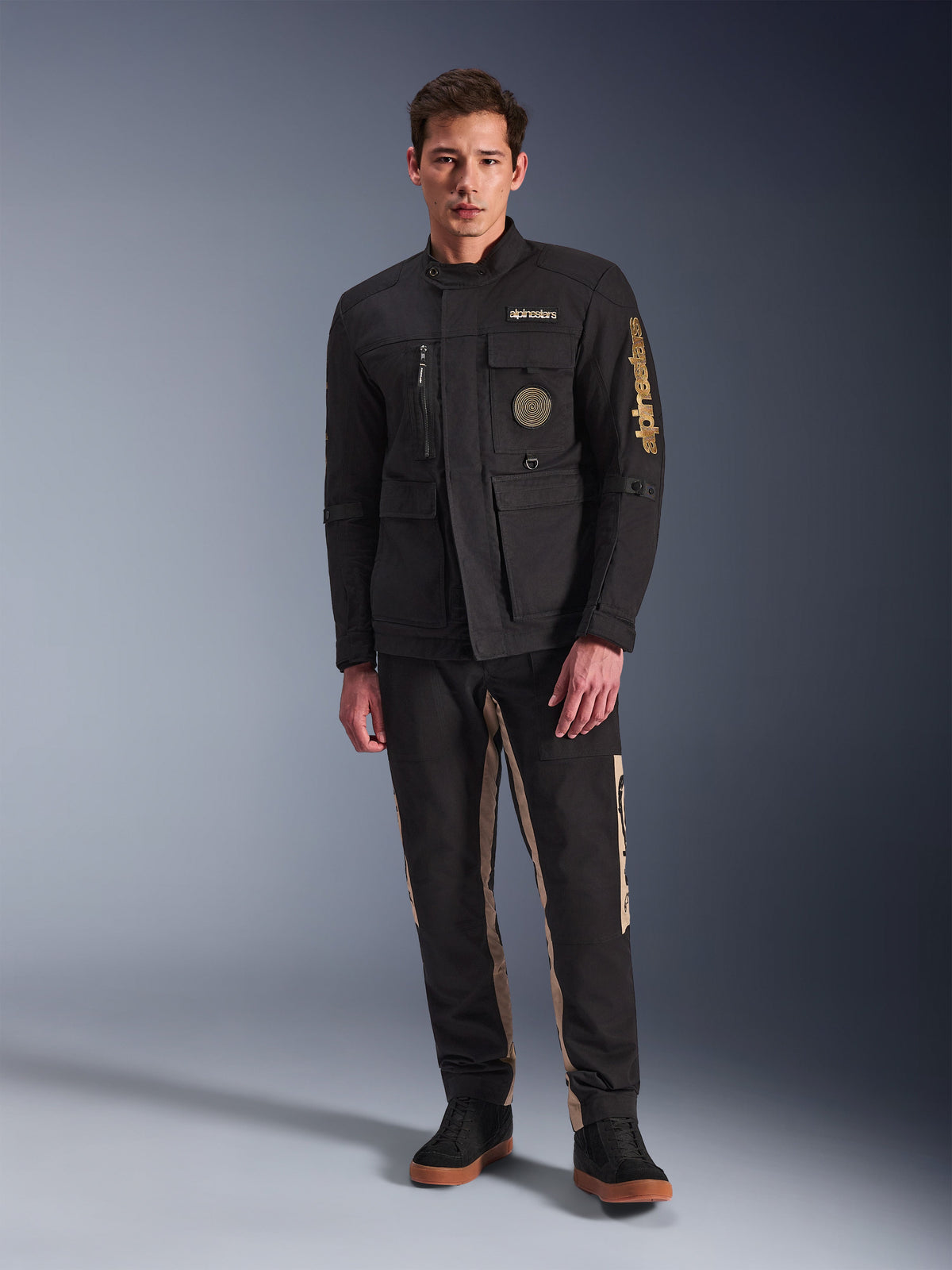 Mandlig model iført Alpinestars Dice Jacket, sort urban motorcykeljakke med vintage feltjakke-stil, fire funktionelle frontlommer, Alpinestars-branding på ærmet og brystet, kombineret med sorte og sandfarvede bukser, moderne urban køreudstyr