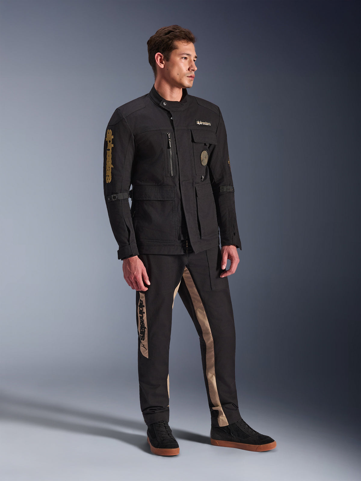 Mandlig model iført Alpinestars Dice-jakke i sort, urban motorcykeljakke med vintage feltjakke-æstetik, med flere funktionelle lommer og guld-branding på ærmet, kombineret med matchende sorte og sandfarvede bukser og sorte sneakers.