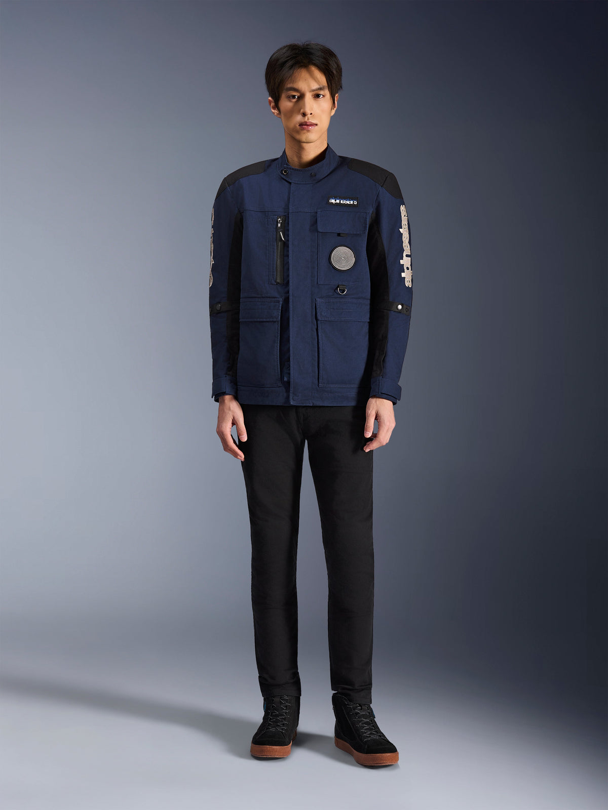 Alpinestars Dice-jakke, urban motorcykeljakke, mørkeblå og sort, bæres af en model med sorte bukser og sorte sko, moderne feltjakke-design med flere lommer, lodret brystlynlås og hvidt Alpinestars-branding på ærmerne.