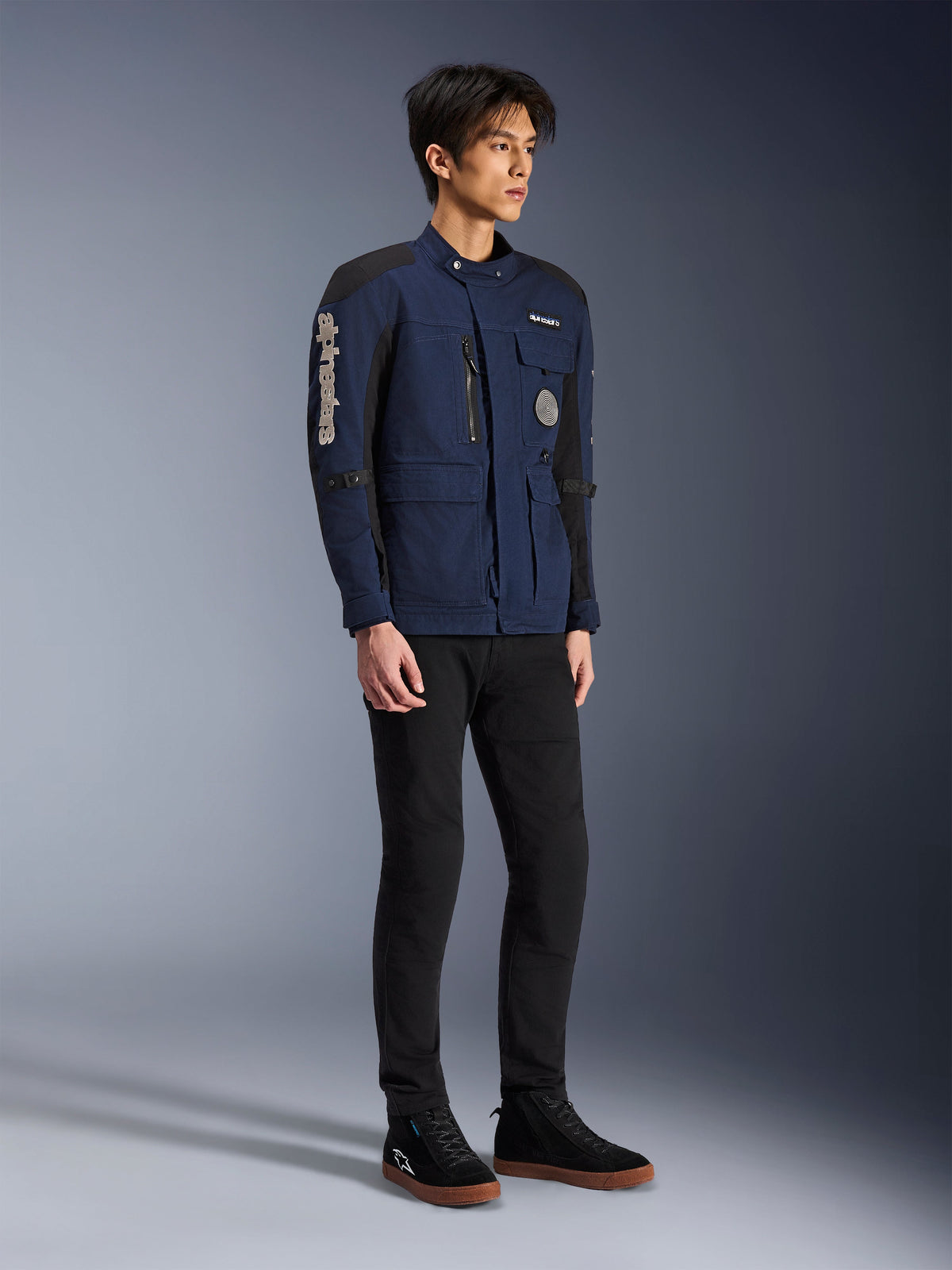 Alpinestars Dice Jakke, mørkeblå og sort, moderne motorcykeljakke i felt-stil båret af en model med sorte bukser og sorte high-top sneakers, med flere praktiske lommer, lodret Alpinestars-branding på ærmet og vævede mærker, urbant moderne køreudstyr