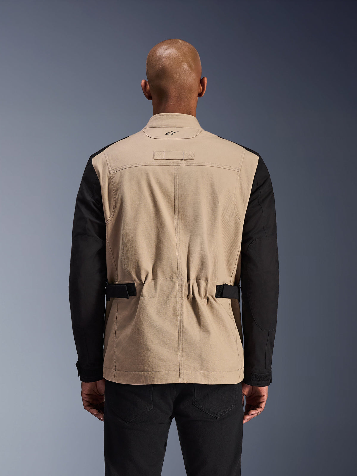 Alpinestars Dice-jakke, urban motorcykeljakke, Aluminum og sort, set bagfra på en model, med plisserede skuldre for mobilitet, justerbare taljeremme og en robust polybomulds-twill-konstruktion.