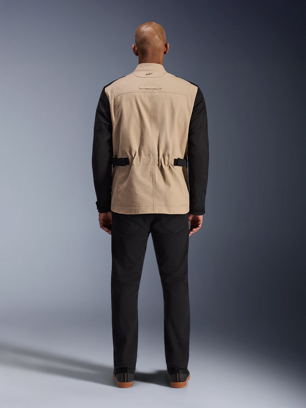 Alpinestars Dice-jakke, urban motorcykeljakke, tan og sort, set bagfra på model, med polybomuld twill-konstruktion, justerbare taljestropper og lille broderet logo på kraven, båret med sorte bukser.